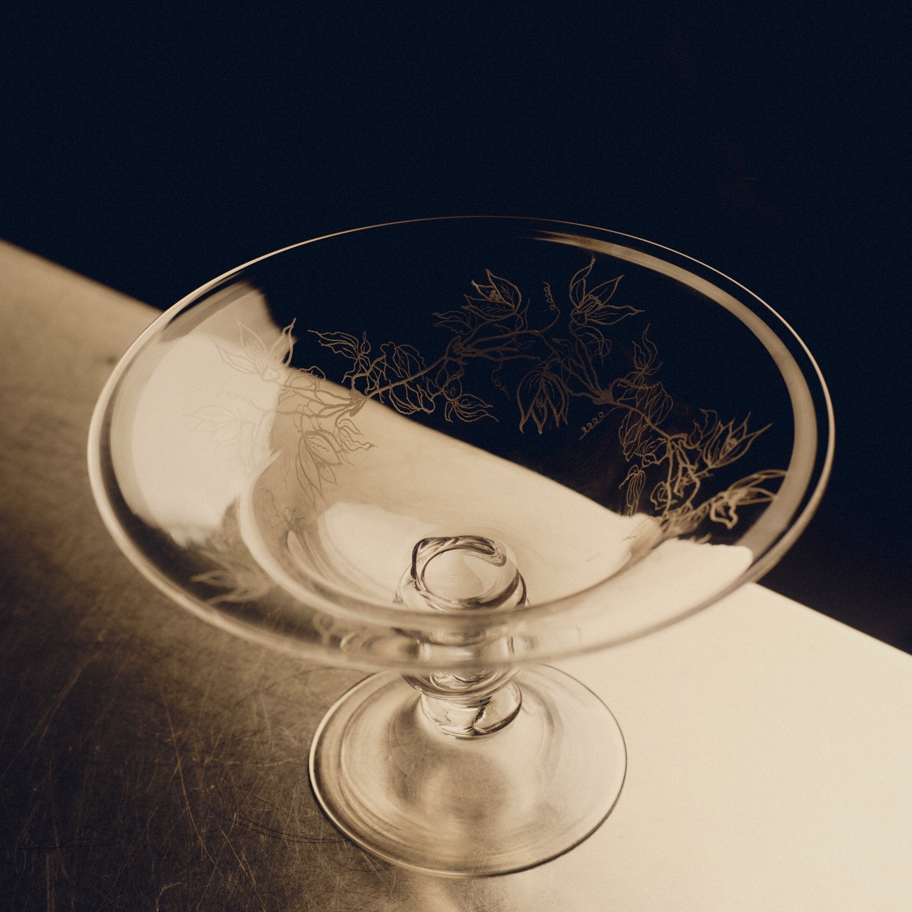Footed glass bowl with floral engraving by Bastian Bjerge sitting on a stainless steel countertop.