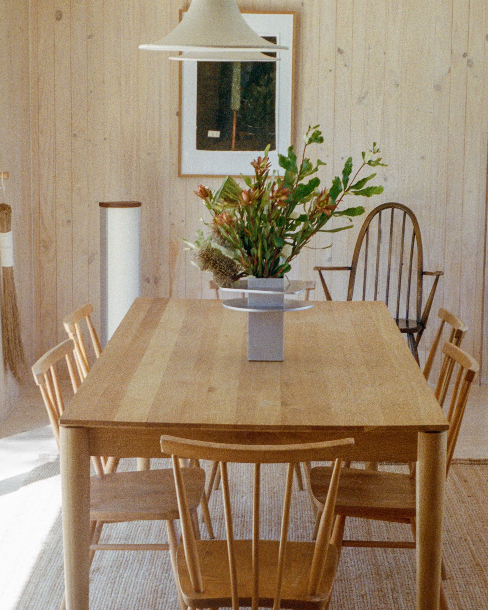 Vase by PARCTA holding tropical flowers on a wooden table in a cabin setting. There are six wooden chairs around the table and above hangs a pendant lamp with a picture on the back wall.