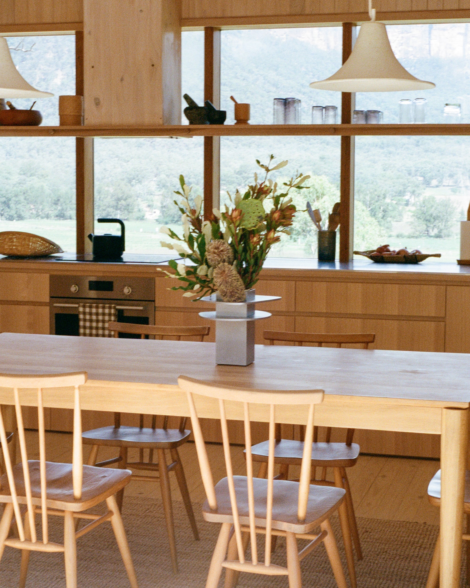 Vase by PARCTA holding tropical flowers on a wooden table in a the kitchen of a cabin. Windows at the back of the kitchen look out on a greenery setting.
