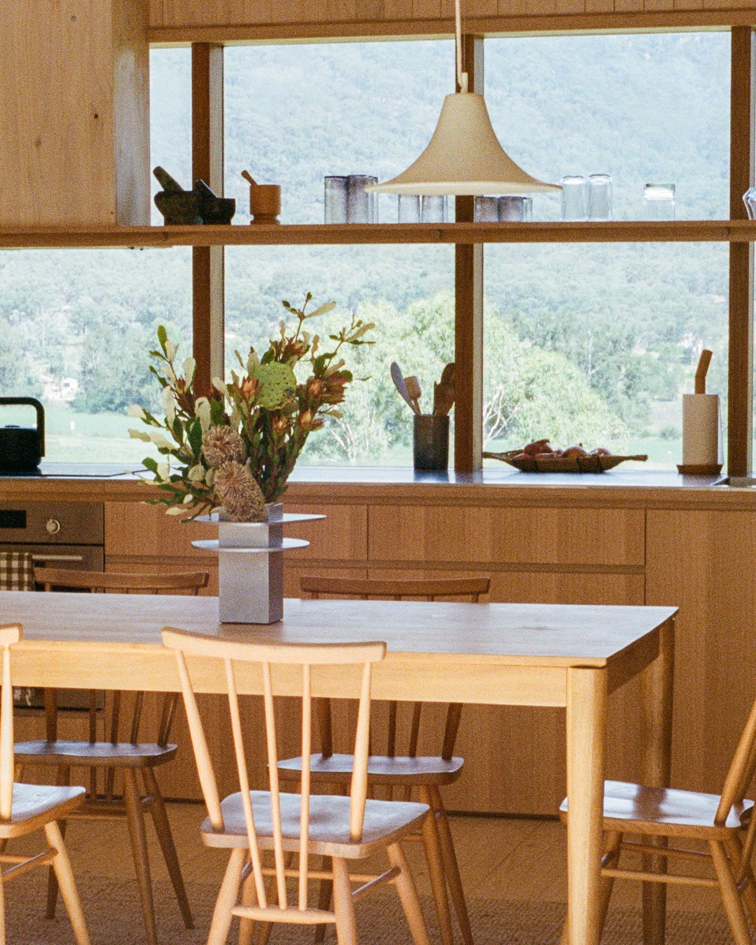 Vase by PARCTA holding tropical flowers on a wooden table in a the kitchen of a cabin. Windows at the back of the kitchen look out on a greenery setting.