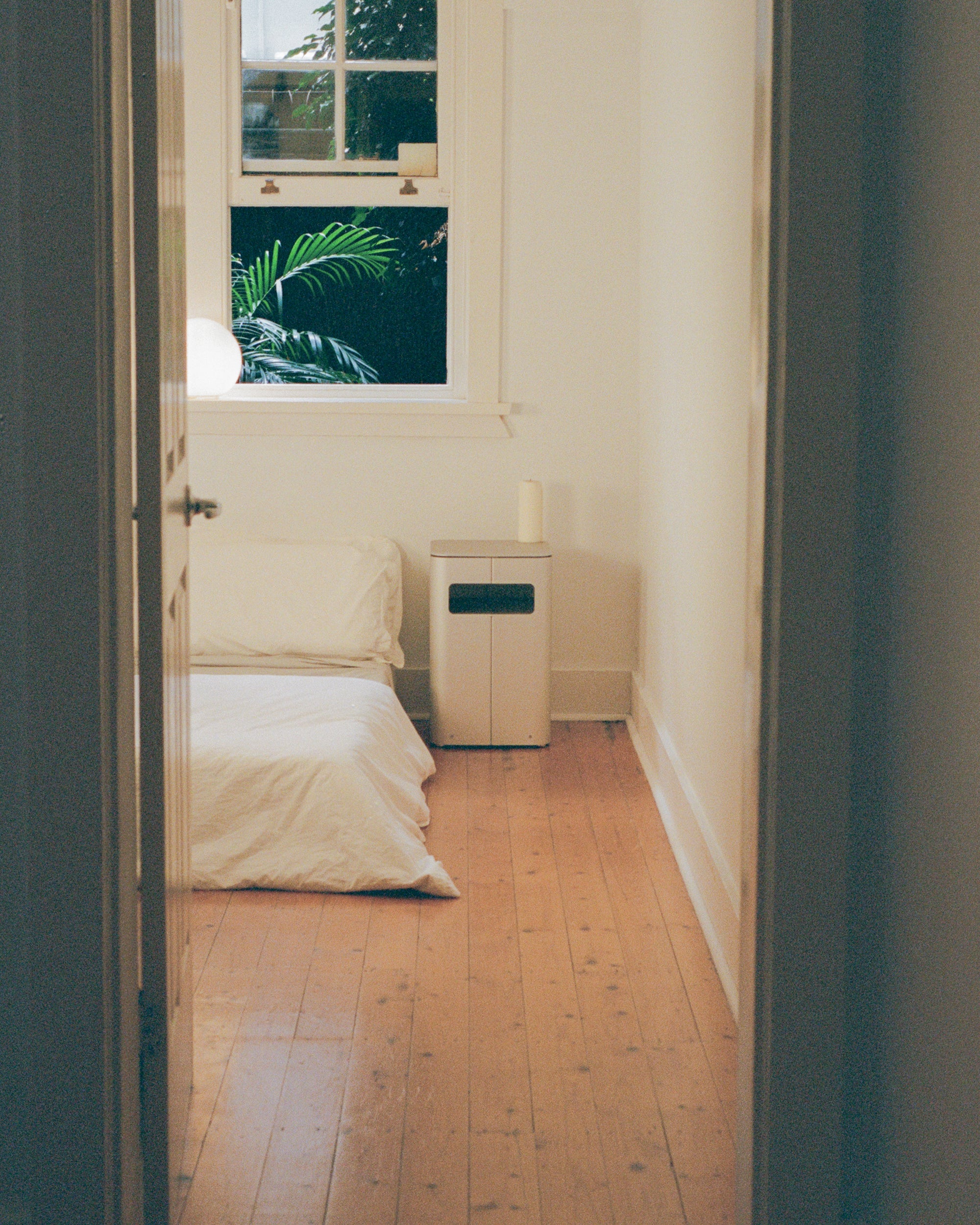 Looking into the bedroom where a bedside table by PARCTA is placed besides a bed with white linen. There's an open window above the bed that's showing leaves of a large plant. 