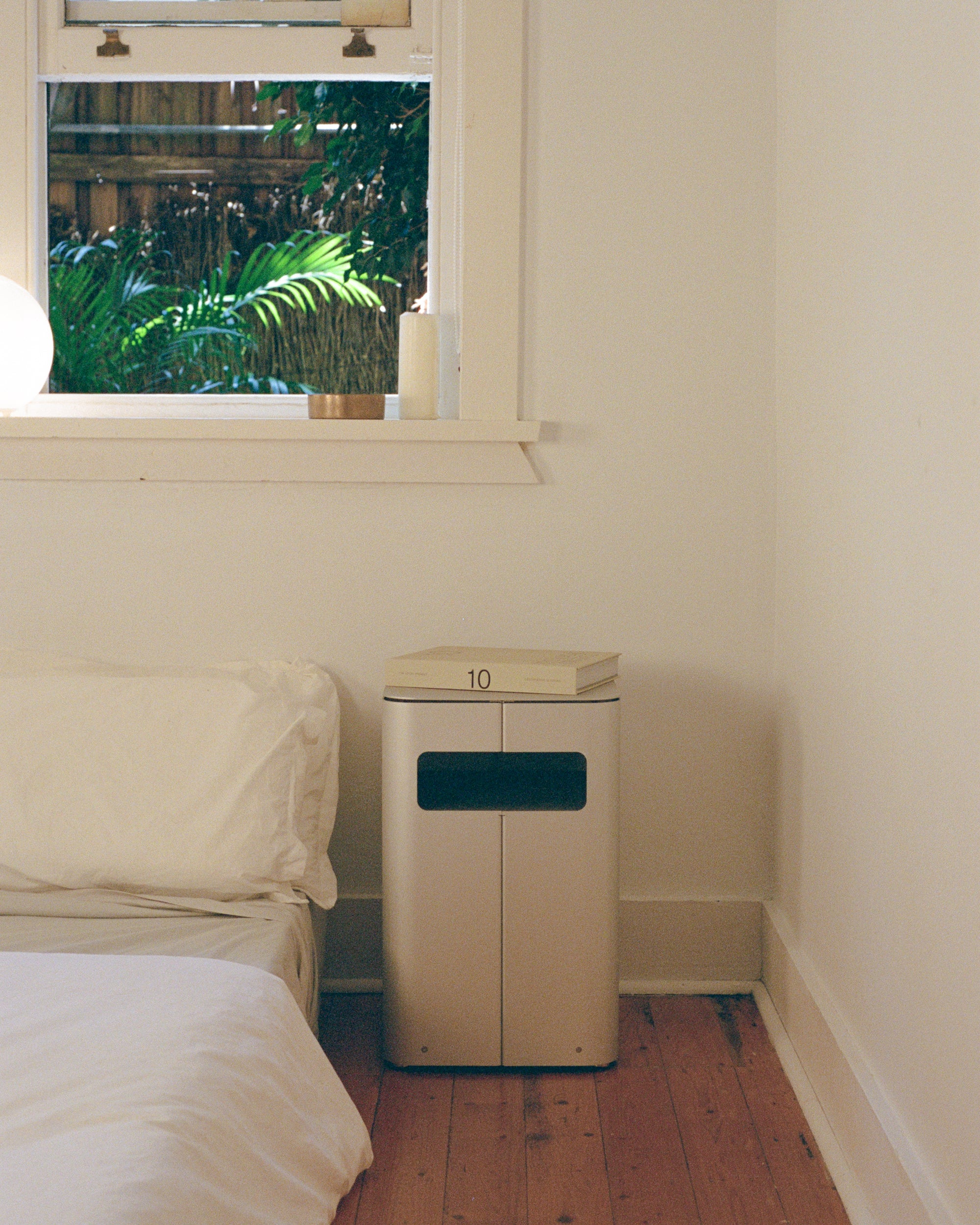 Bedside table by PARCTA placed besides a bed with white linen. There's an open window above the bed that's showing leaves of a large plant.
