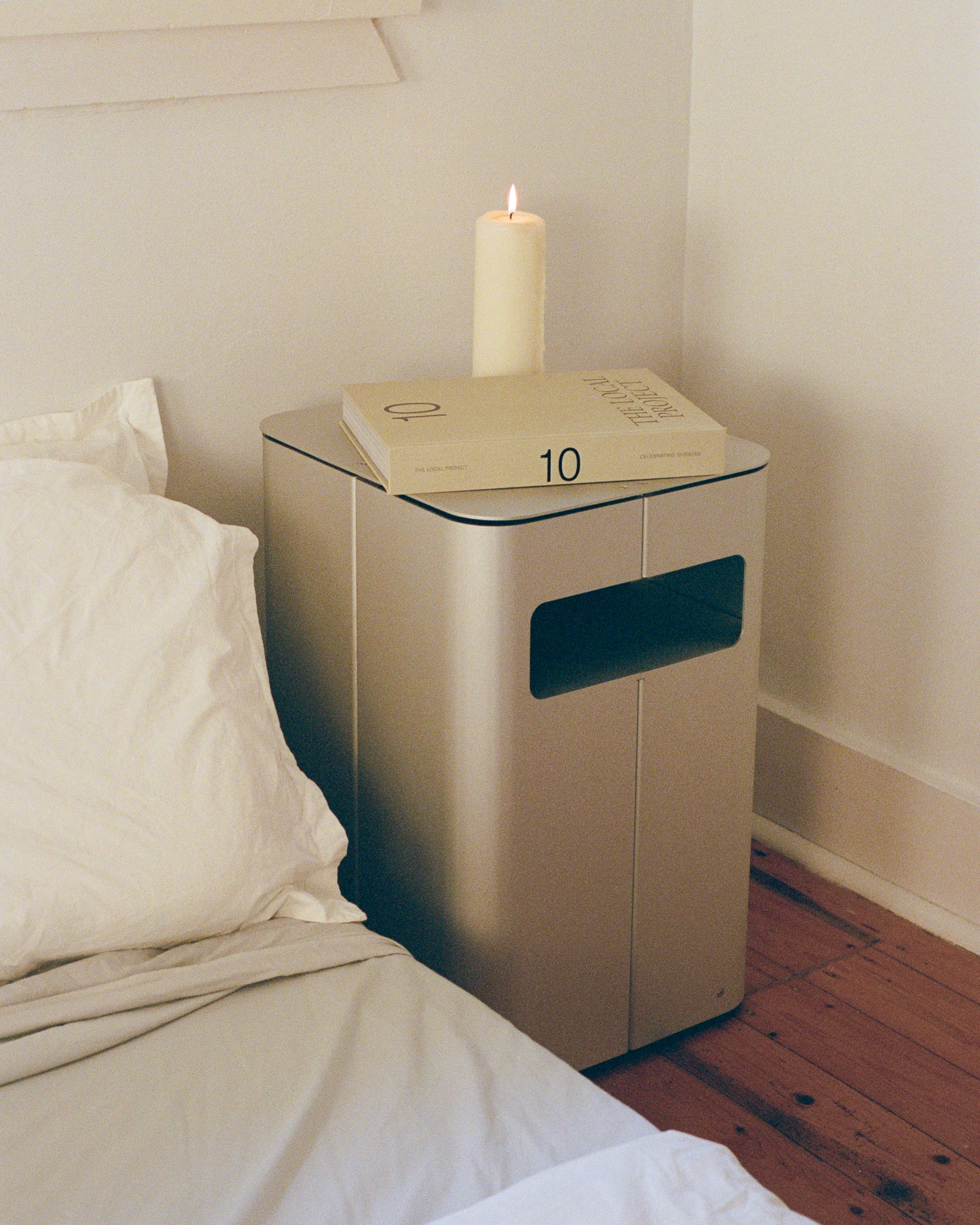 Bedside table by PARCTA placed besides a bed with white linen. On top of the bedside table is a candle and a book.