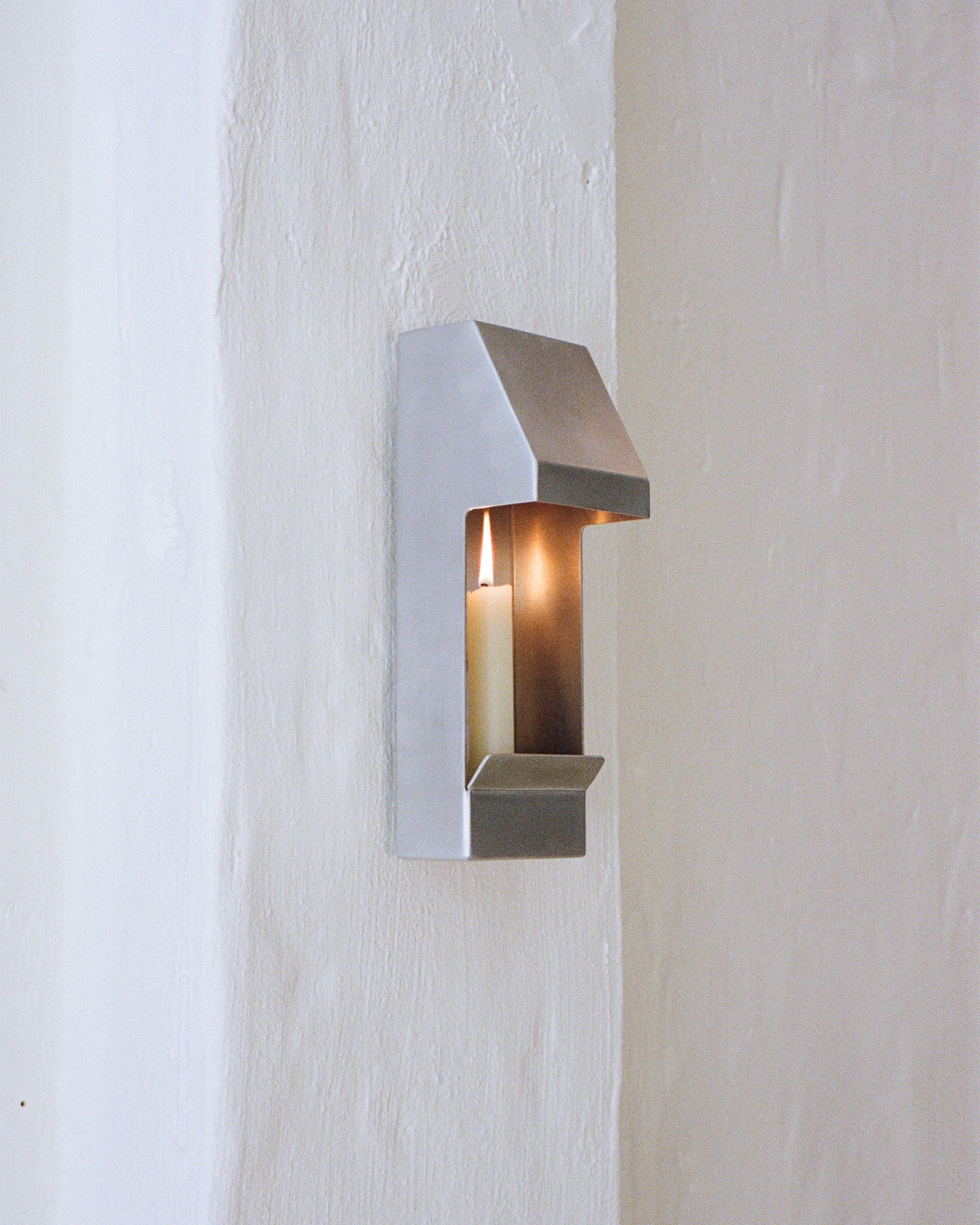 Candle holder by PARCTA with a lit candle inside on a white painted, textured wall.