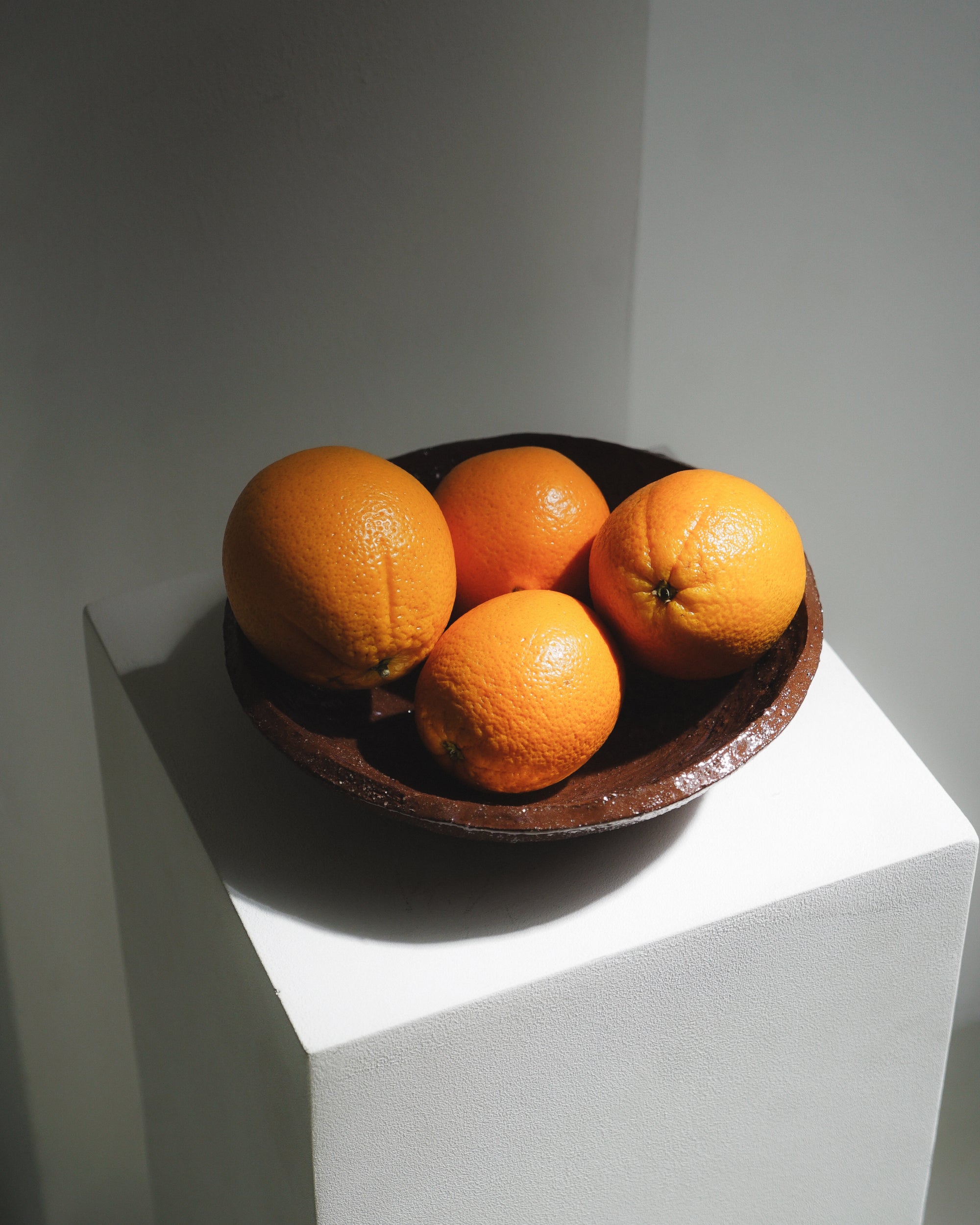 Terracotta bowl by klangnapha finished with a shiny, transparent glaze that's holding oranges and sat on top of a white plinth on a white background.