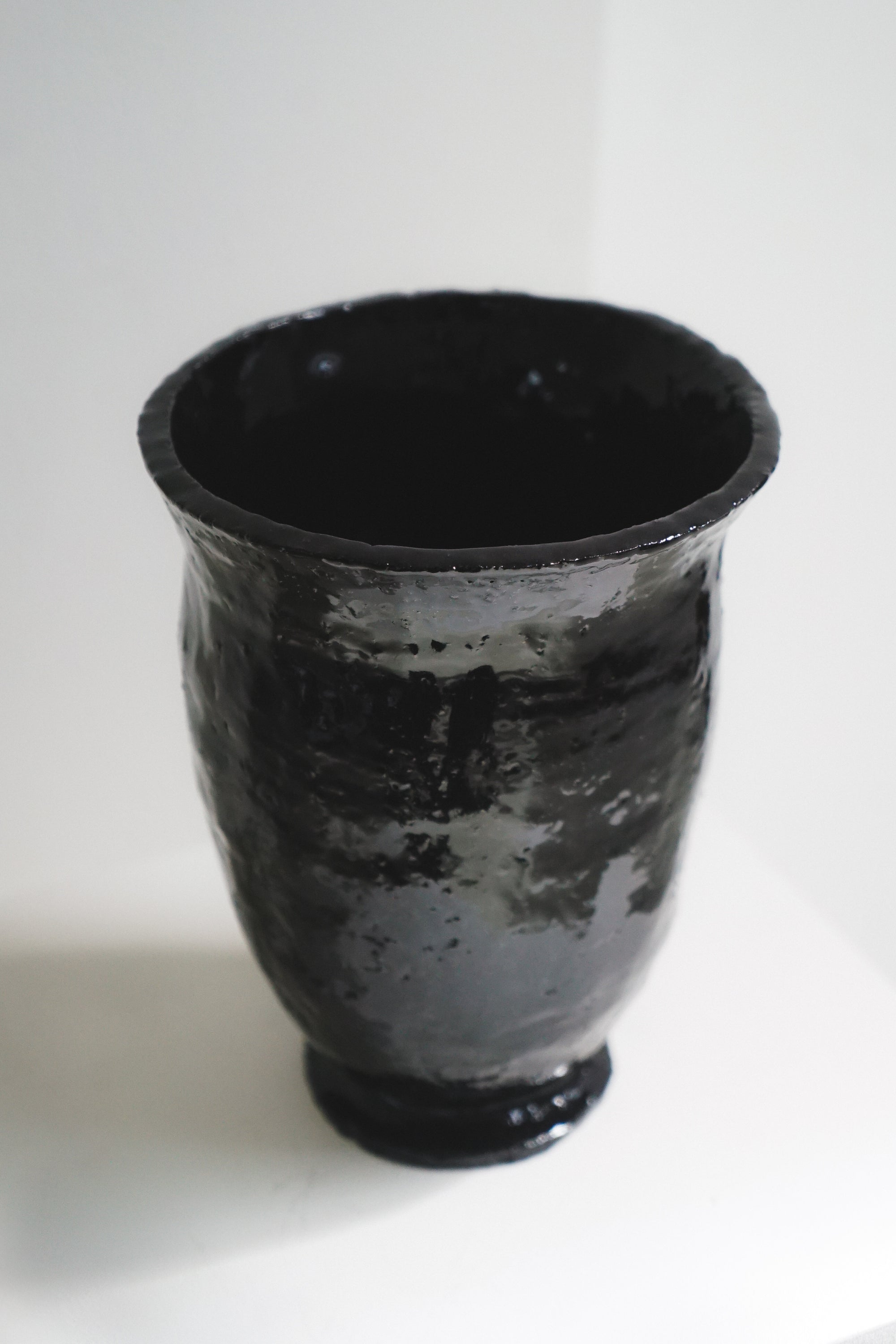 Black, glossy ceramic chalice vase by klangnapha on a white plinth with a neutral background.