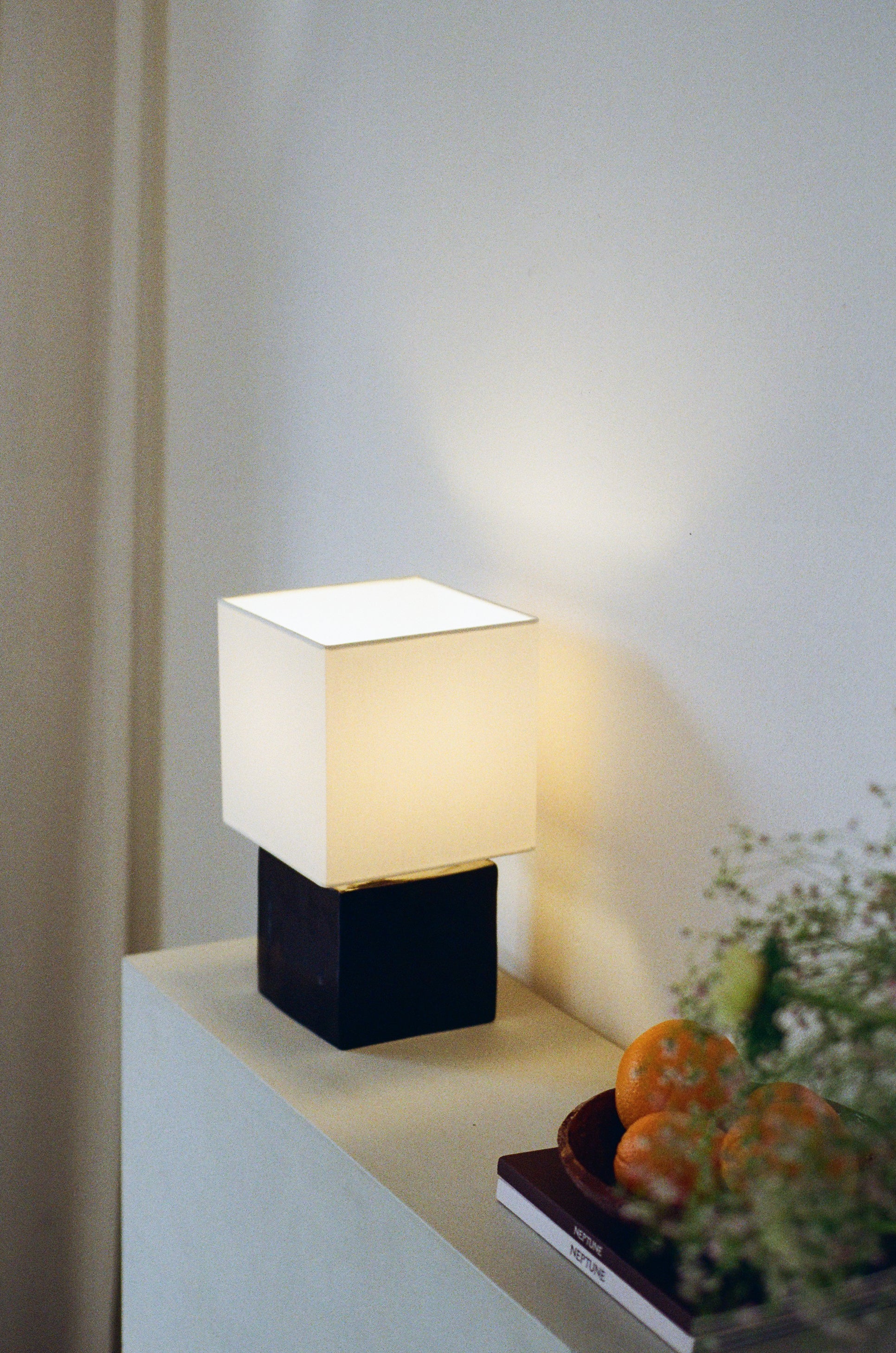 Cream geometric table lamp with light on by klangnapha with black, glossy cube base sat on a wall, besides a fruit bowl, books and slight greenery.