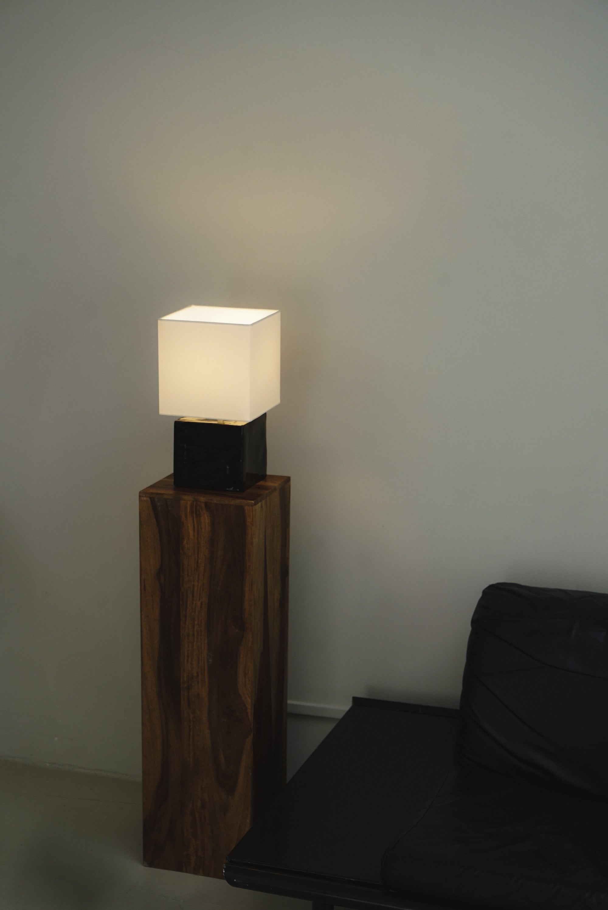Cream geometric table lamp with light on by klangnapha with black, glossy cube base on wooden plinth with neutral background, besides a black leather sofa.