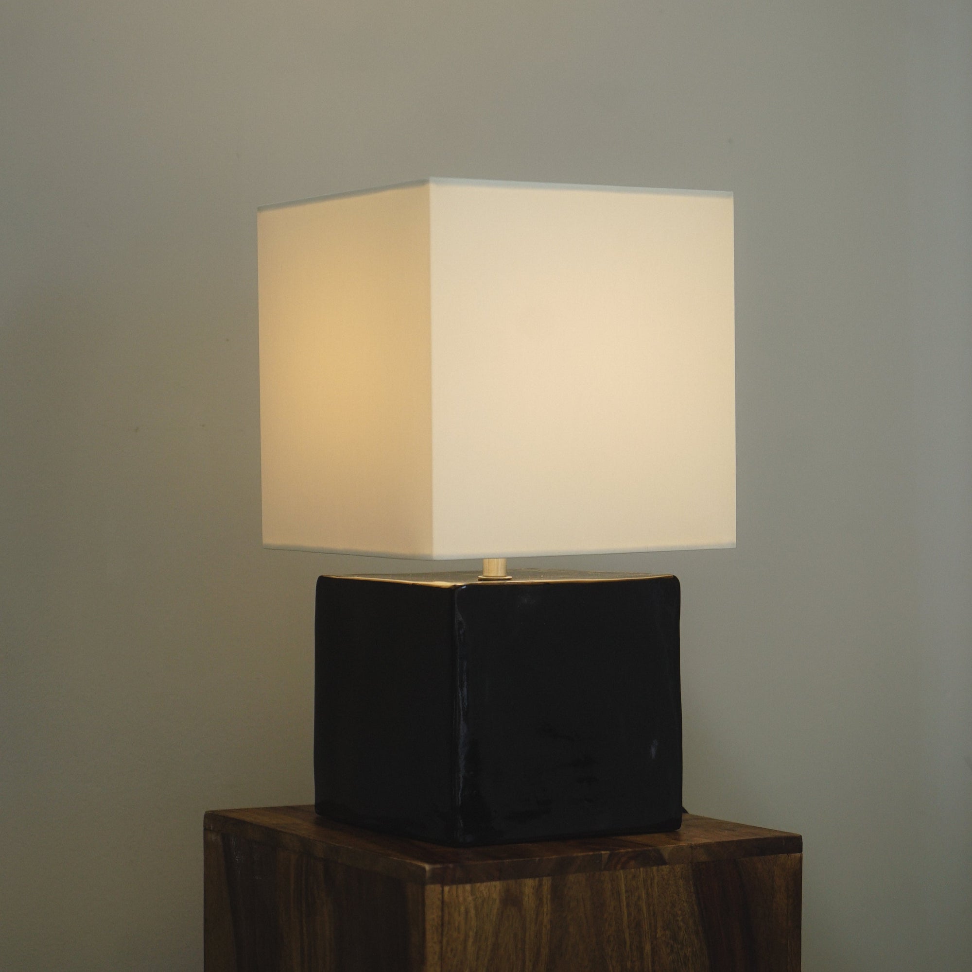 Cream geometric table lamp with light on by klangnapha with a black, glossy cube base on a wooden plinth with a neutral background. 