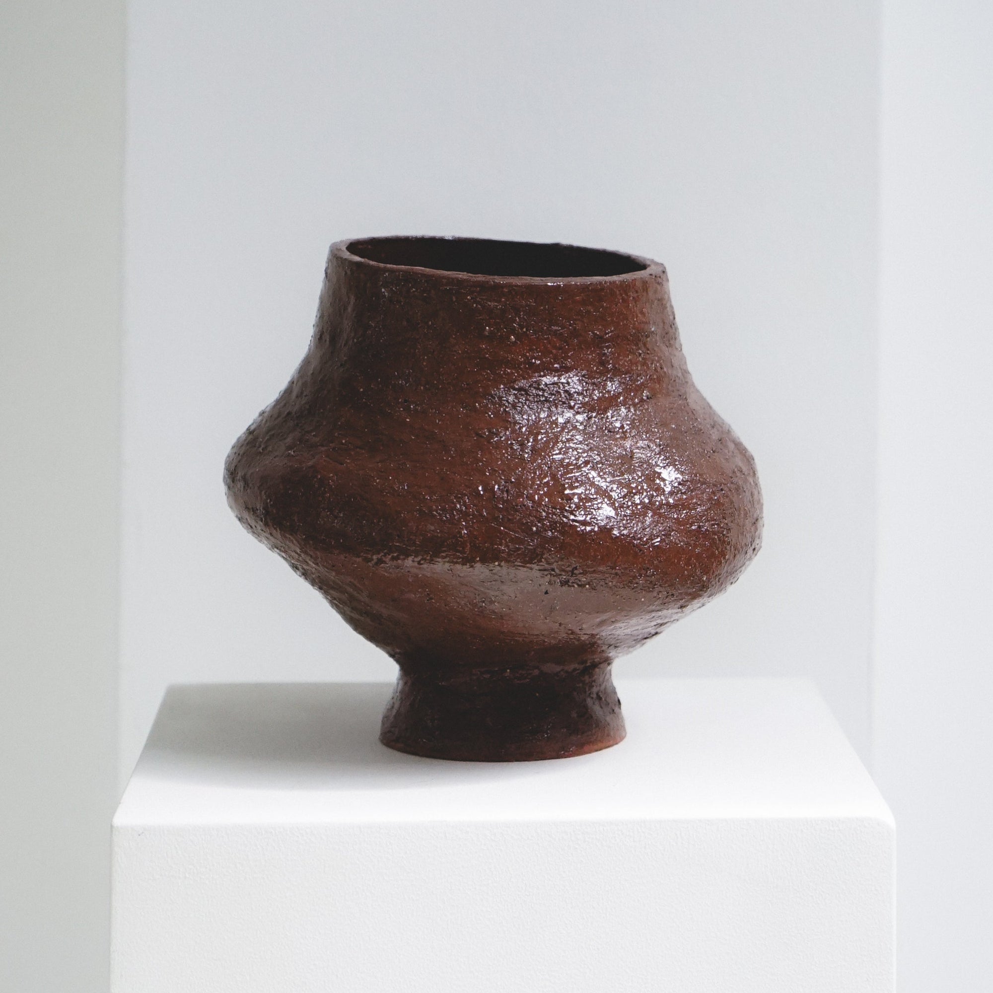 Rounded, brown ceramic vase by klangnapha on a white plinth with a neutral background.