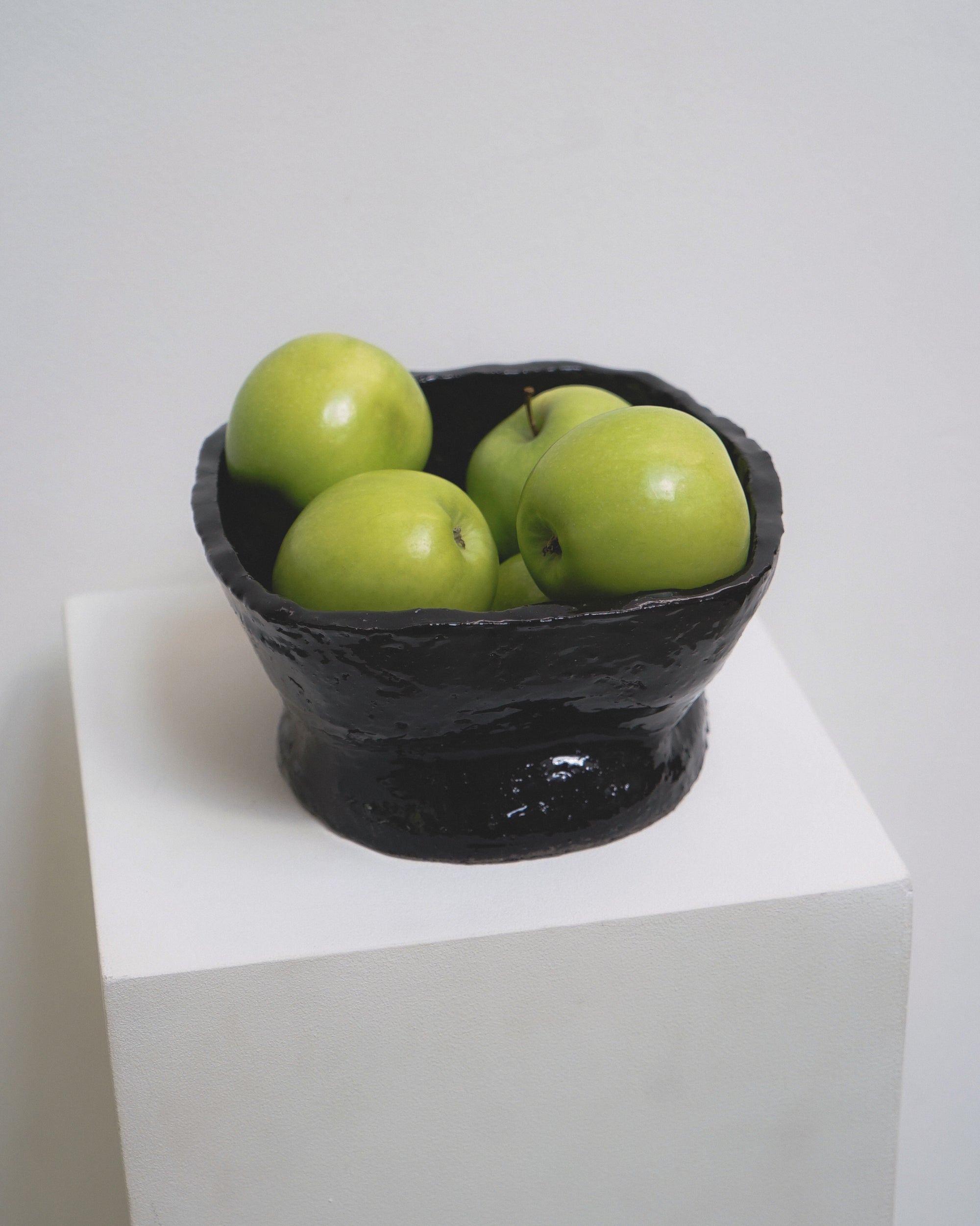 Square ceramic bowl by klangnapha in a dark gloss, sat on top of a white plinth on a white background holding green apples.
