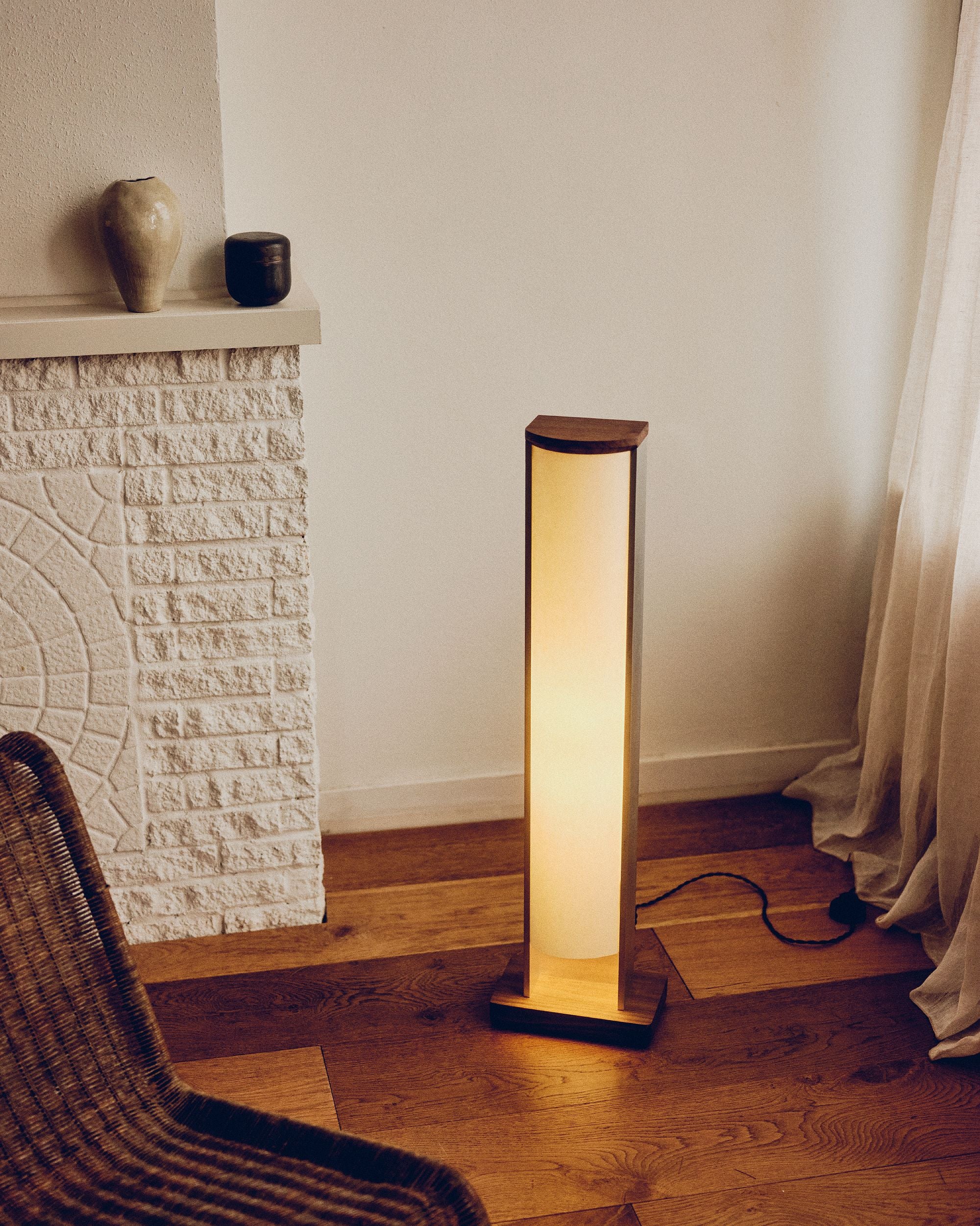 Floor lamp by PARCTA casting a warm glow in a room with a stone fireplace and ceramic objects on top. There's a beige curtain flowing over the wooden floor and the side of a woven chair.