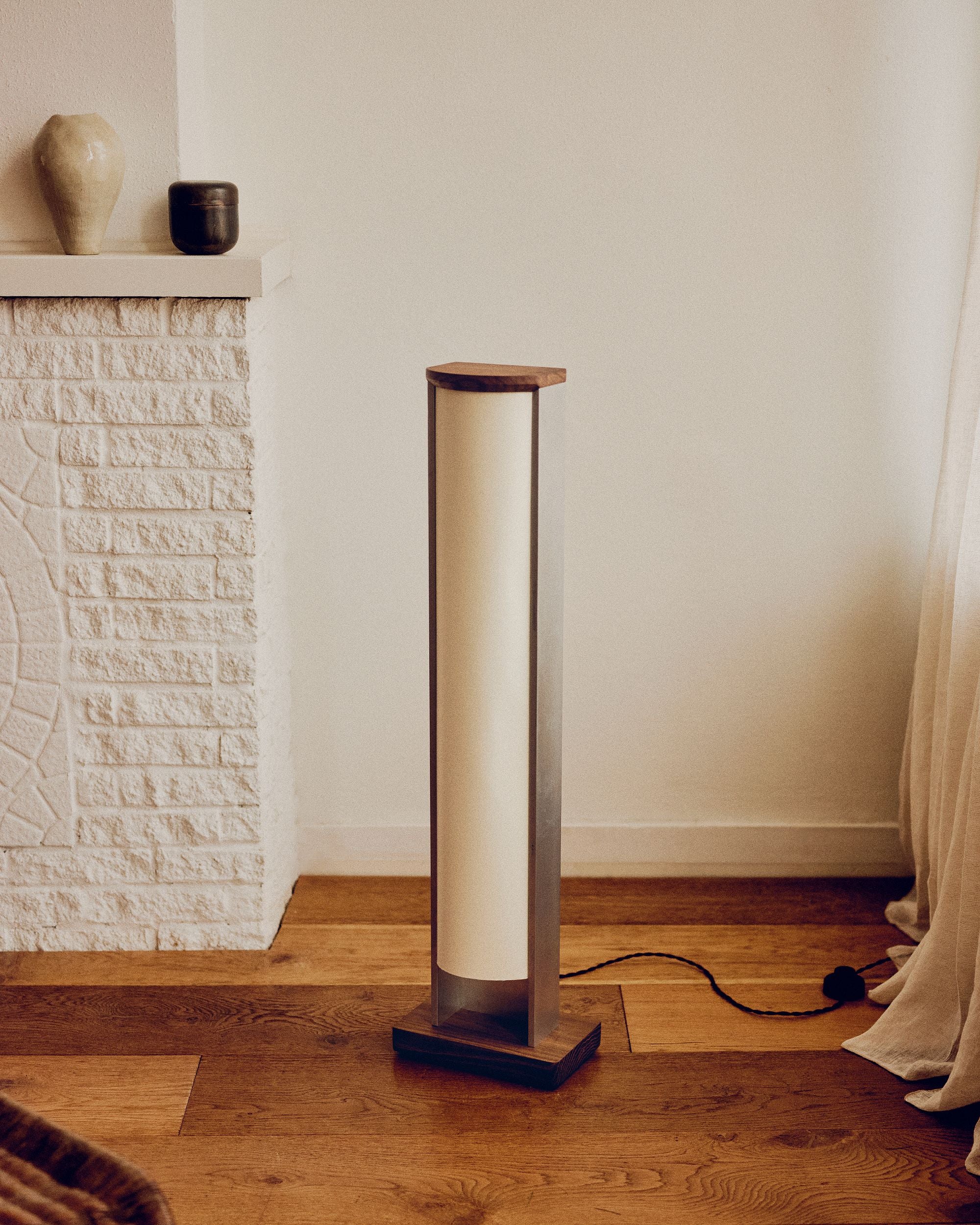 Floor lamp by PARCTA with the light off in a room with a stone fireplace and ceramic objects on top. There's a beige curtain flowing over the wooden floor.