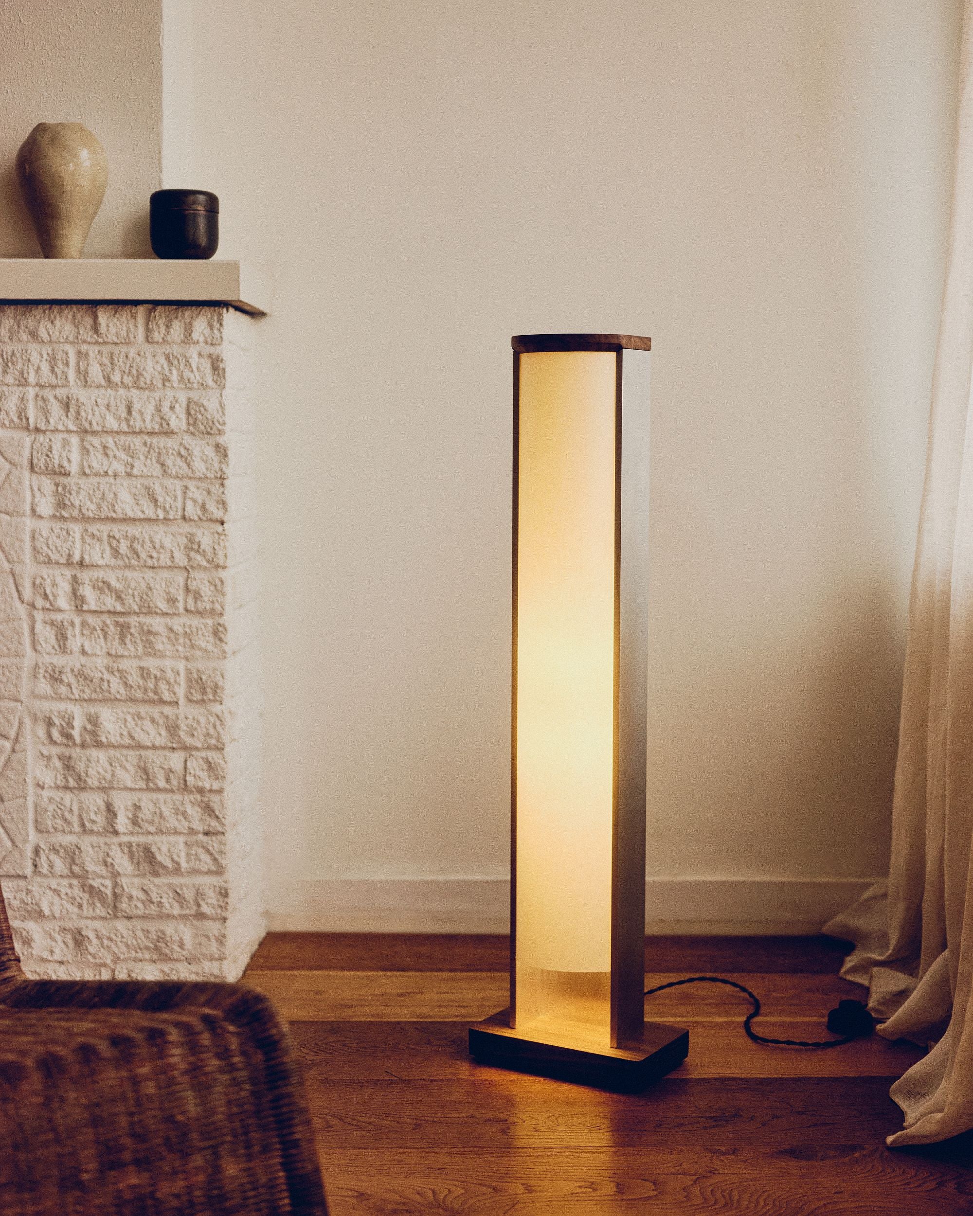 Floor lamp by PARCTA casting a warm glow in a room with a stone fireplace and ceramic objects on top. There's a beige curtain flowing over the wooden floor.