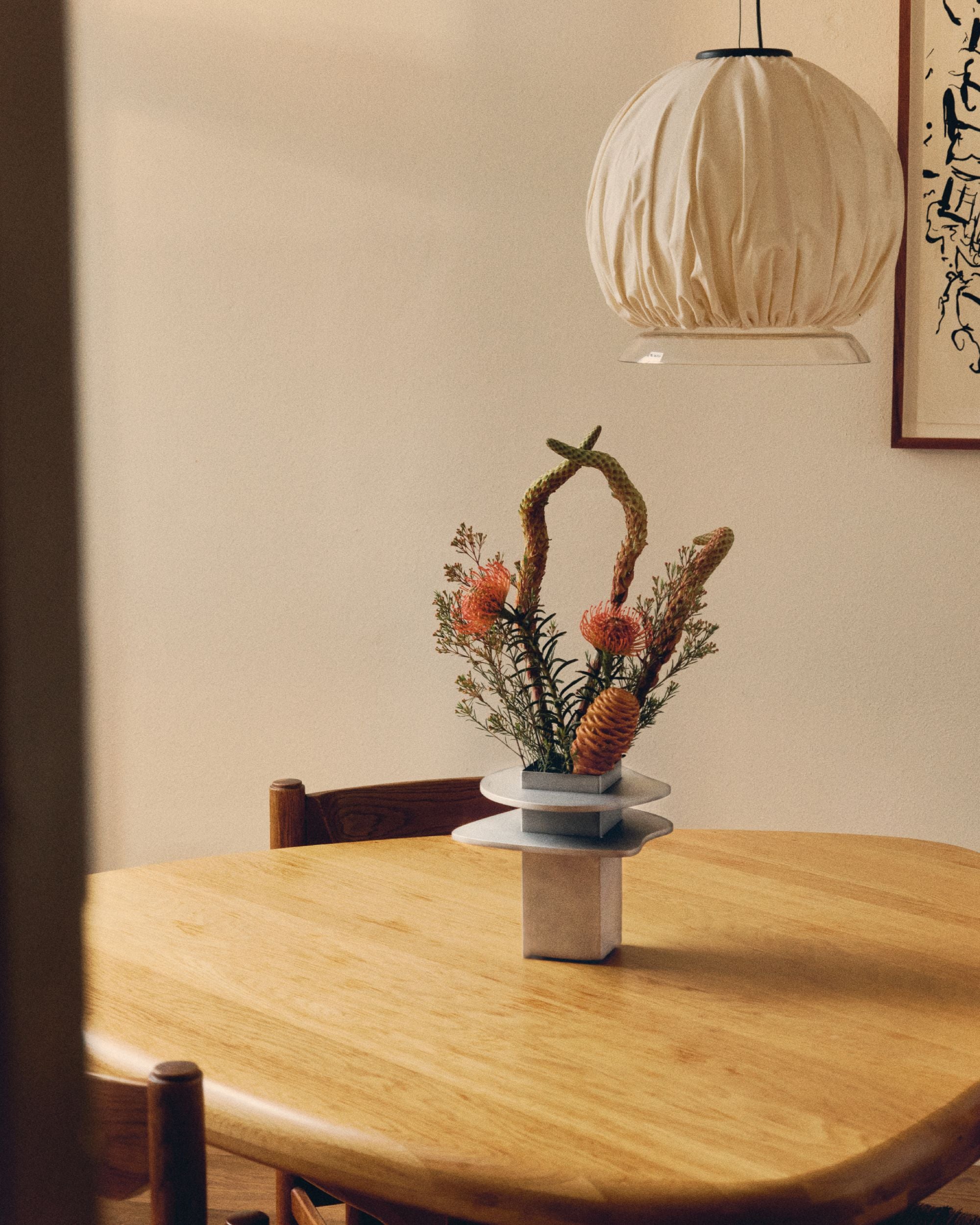 Vase by PARCTA on wooden table with tropical flowers. Above is a beige hanging light and besides that there is the left side of a painting.