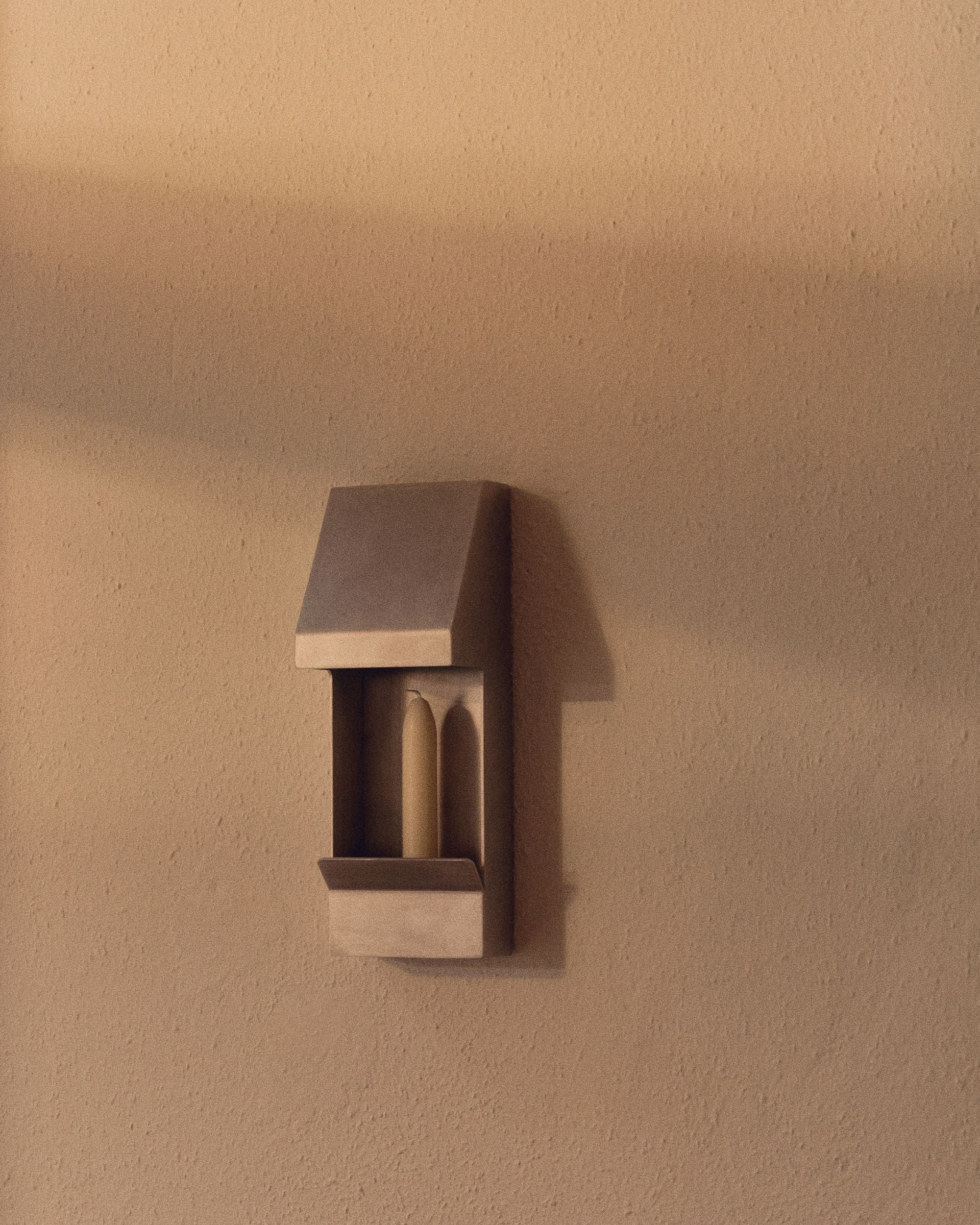 Candle holder by PARCTA with an unlit candle inside on a neutral wall with sunlight shining against it.
