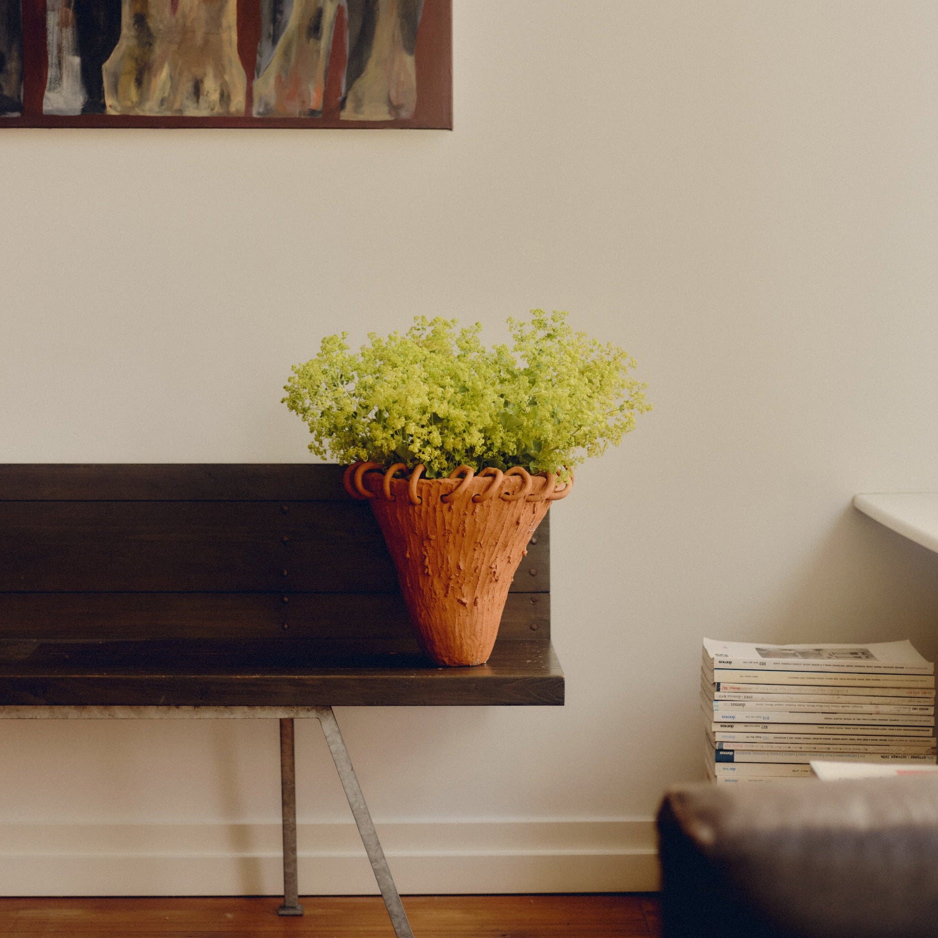 Terra cotta vase with a border of terra cotta rings by May Liok sat on a wooden bench, holding green flowers. Wall art hangs above the vase and besides is a stack of magazines.