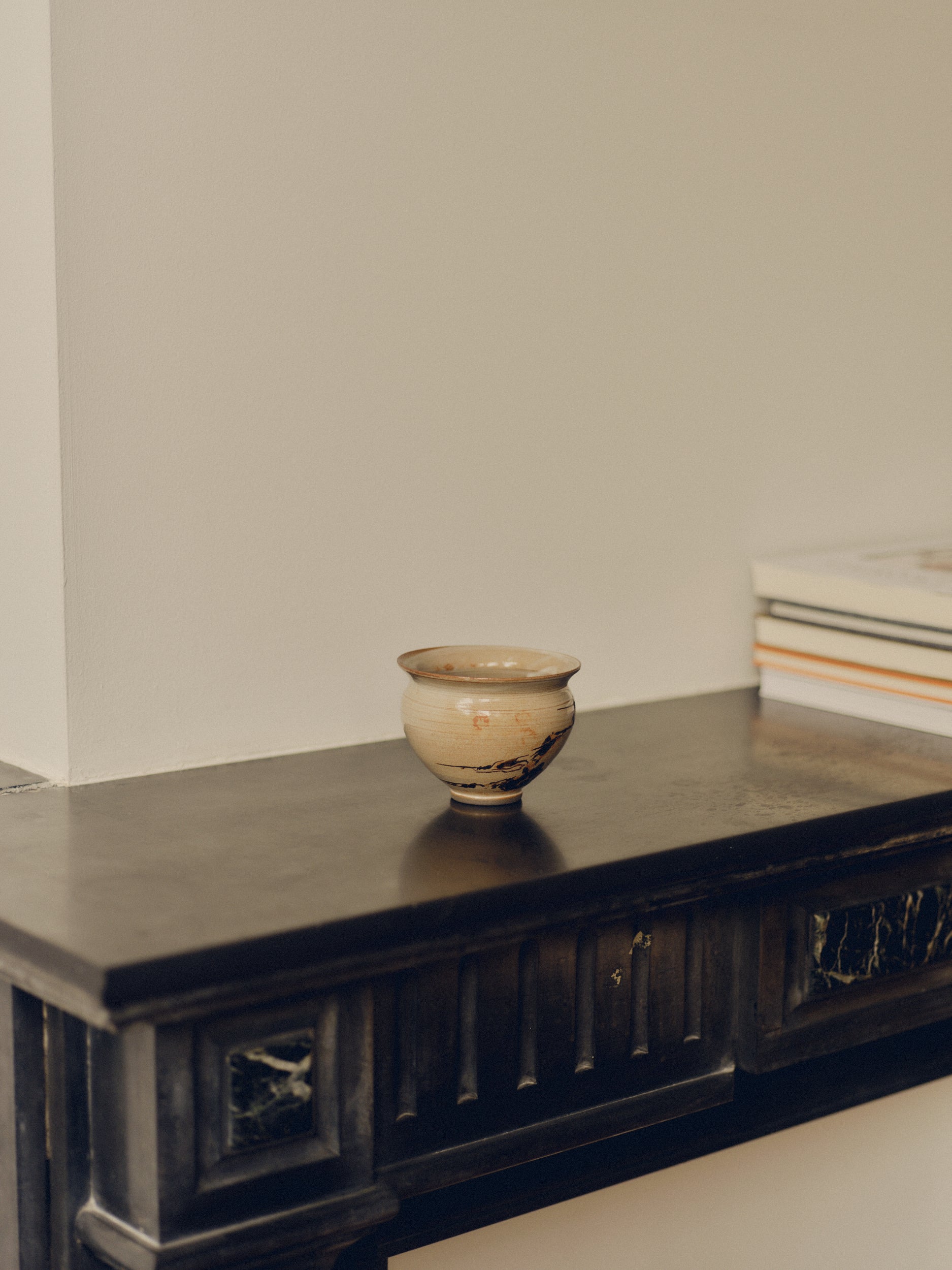 Cream ceramic decorative bowl by Bastian Bjerge with brown hand painted design on a fire place surface, sat besides books.
