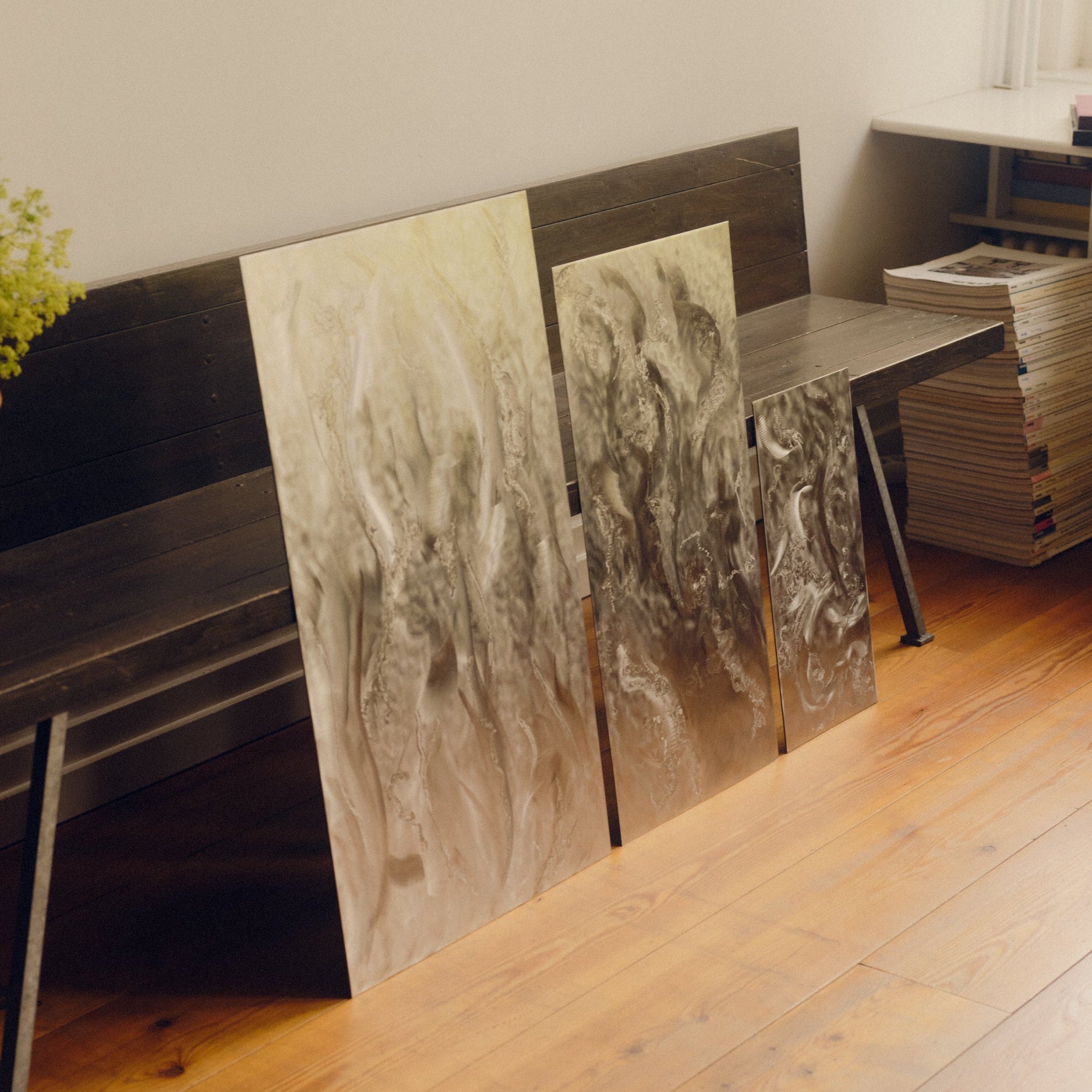 Engraved stainless steel wall art by Grace Houghton, in sizes small, medium and large, leaning against a wooden bench besides a stack of magazines, on a wooden floor.
