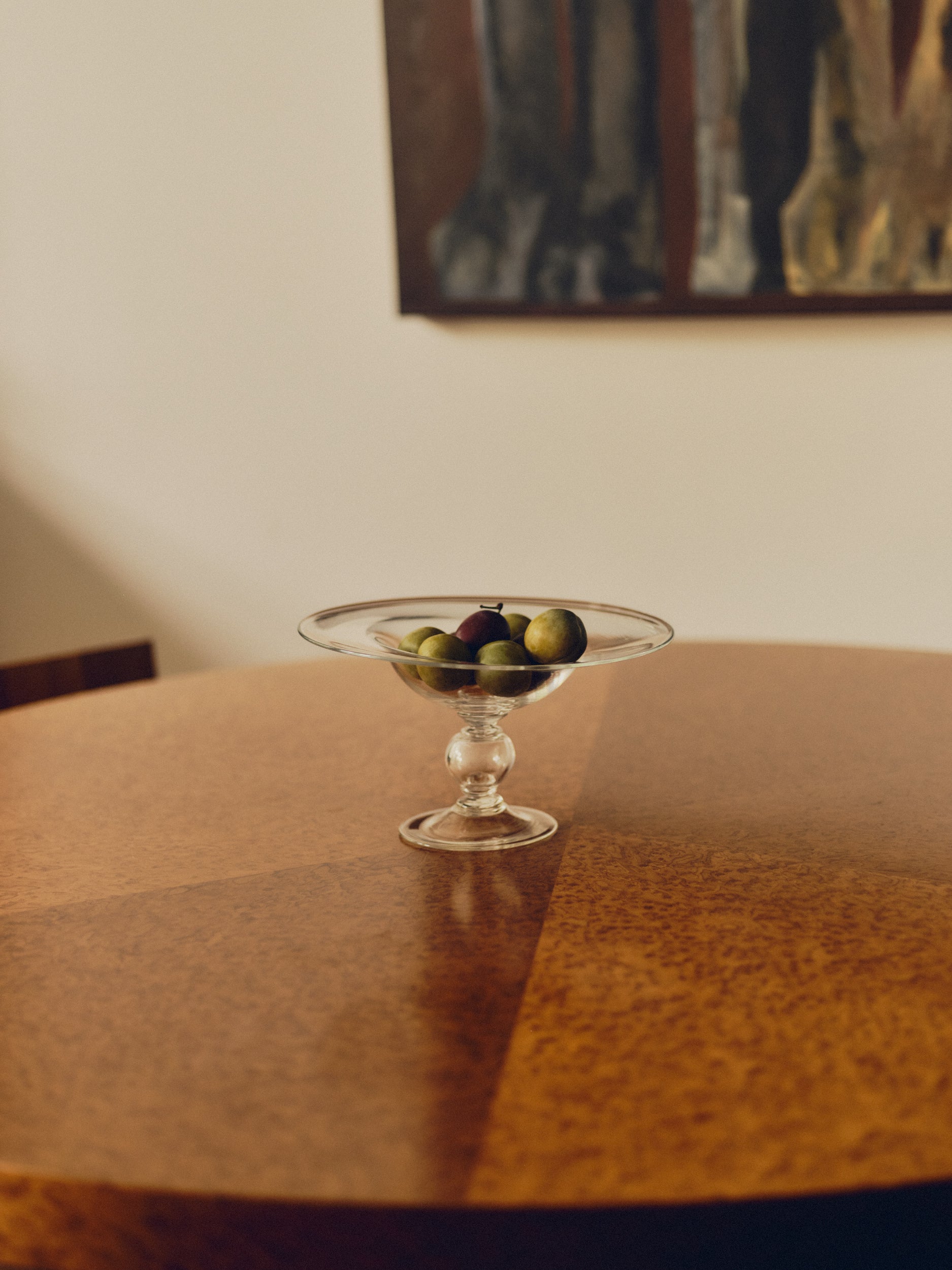 Footed glass bowl by Bastian Bjerge filled with purple and green plums sat on a burl wood table with a painting hanging above. 