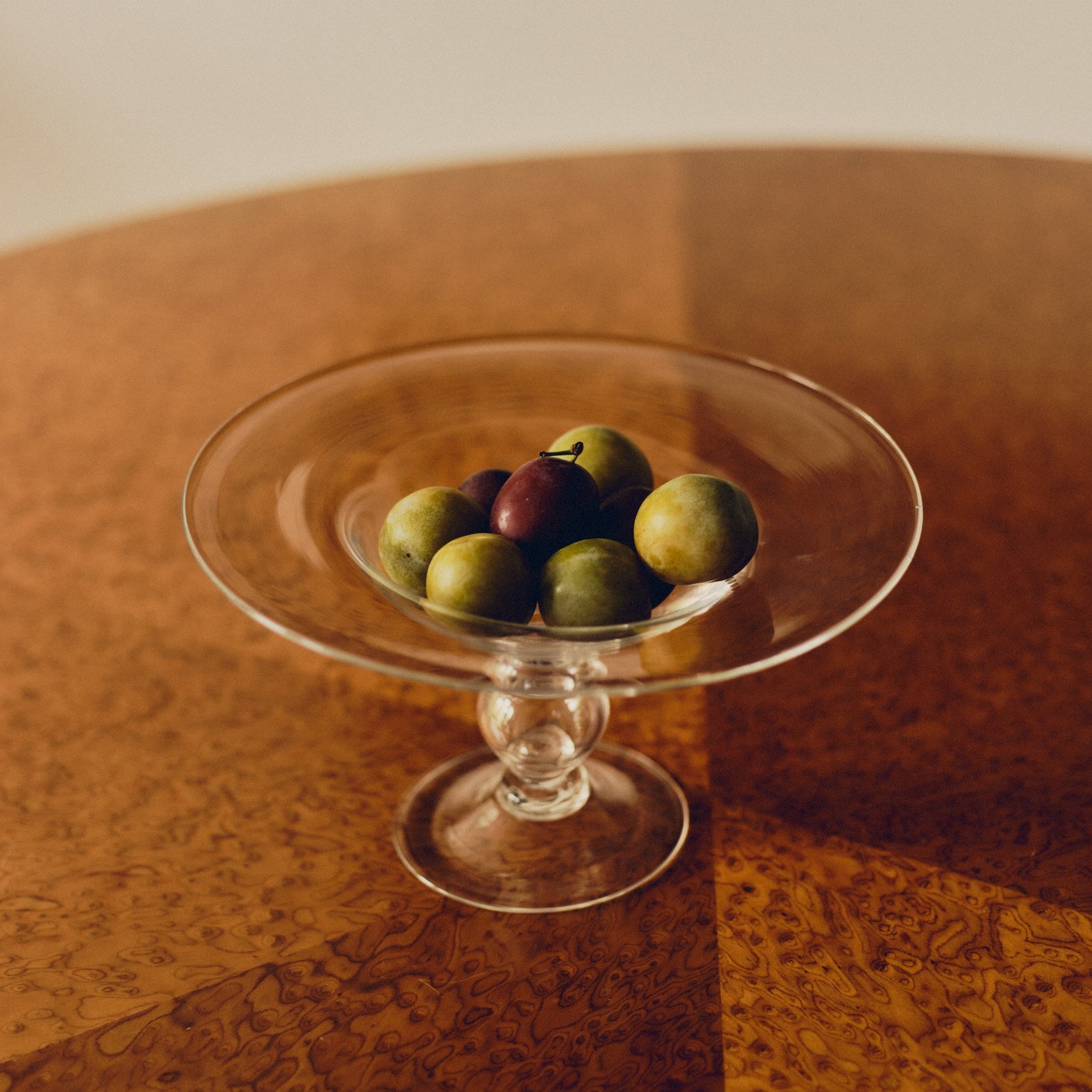 Footed glass bowl by Bastian Bjerge filled with purple and green plums sat on a burl wood table.