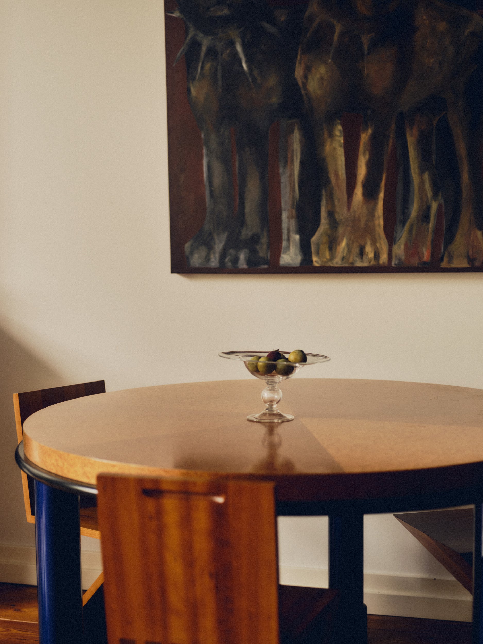 Footed glass bowl by Bastian Bjerge filled with purple and green plums sat on a burl wood table, with Zig Zag chairs underneath and a painting hanging above. 