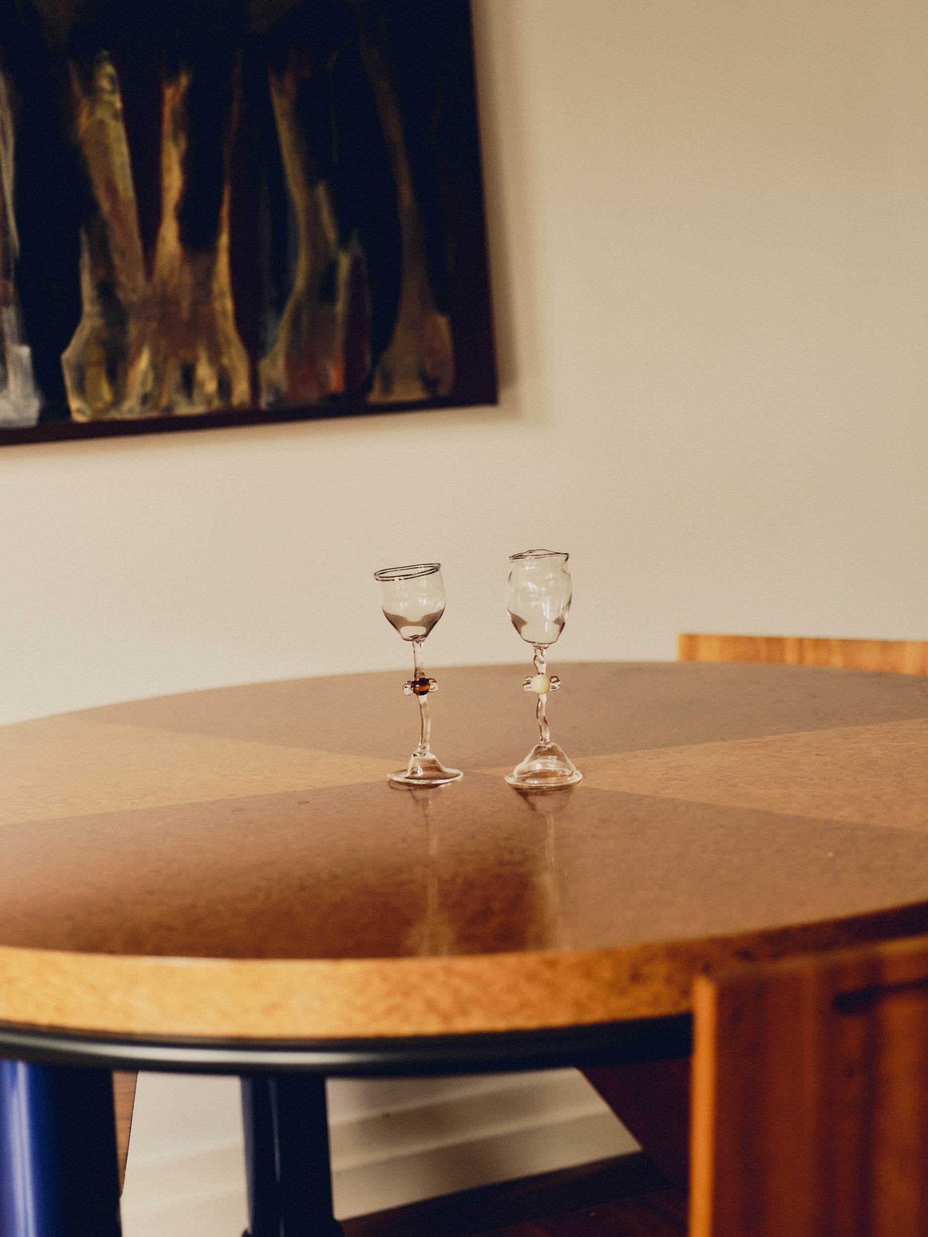 Two mouth-blown wine glasses by Vivian Alarcón. Each wine glass has a candy shaped embellishment on the stem, one is brown and one is ivory. Both glasses are sat on a round burl wood table with a painting hung above.