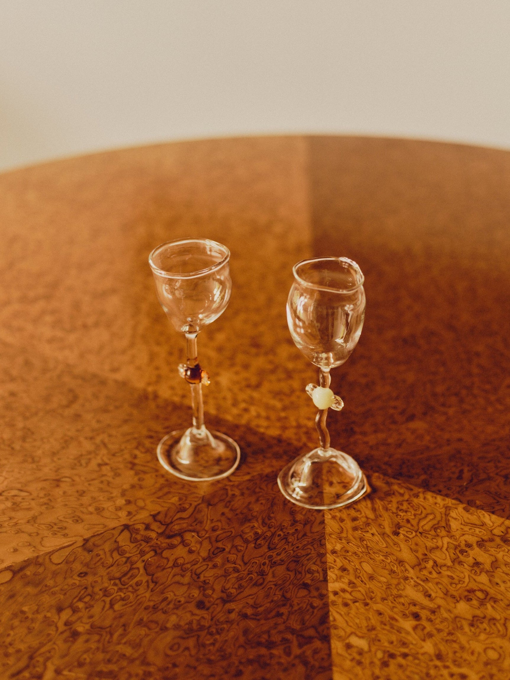 Two mouth-blown wine glasses by Vivian Alarcón. Each wine glass has a candy shaped embellishment on the stem, one is brown and one is ivory. Both glasses are sat on a round burl wood table.