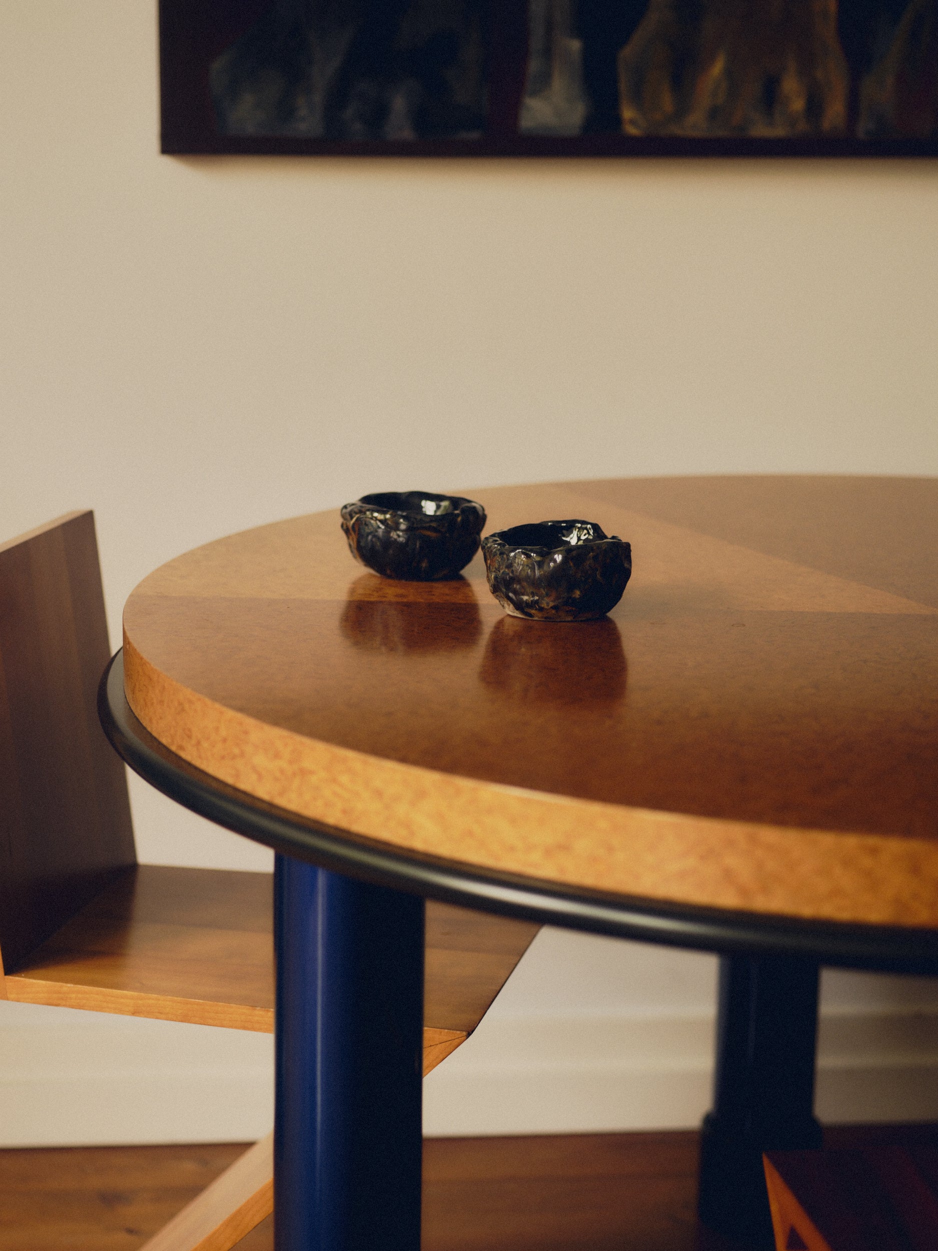 Two textured dark glazed decorative bowls by May Liok sat on a round burl wood table with blue table legs and a Zig Zag chair sat beneath it. Wall art is hanging above the table.