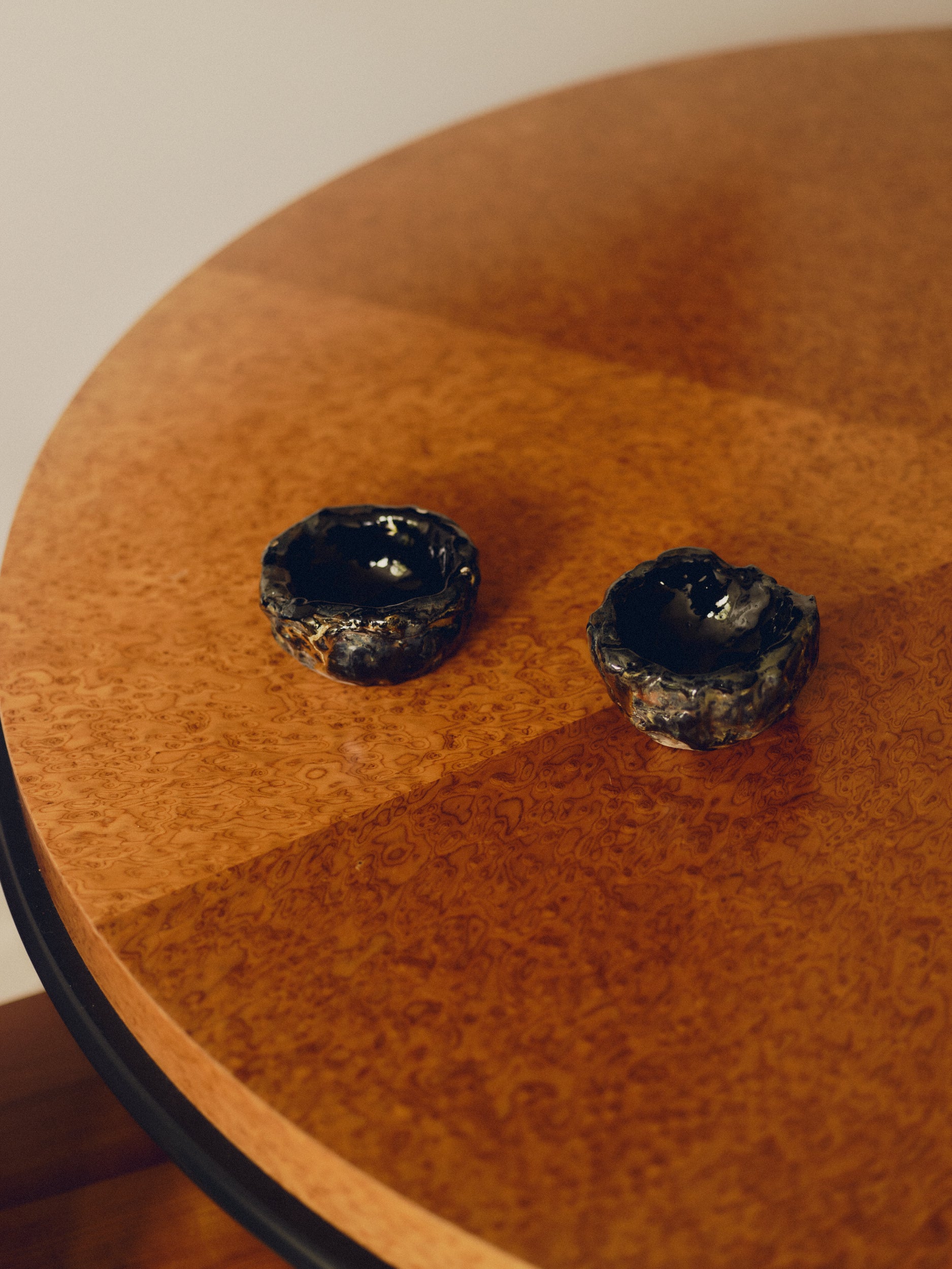 Two textured dark glazed decorative bowls by May Liok sat on a round burl wood table.