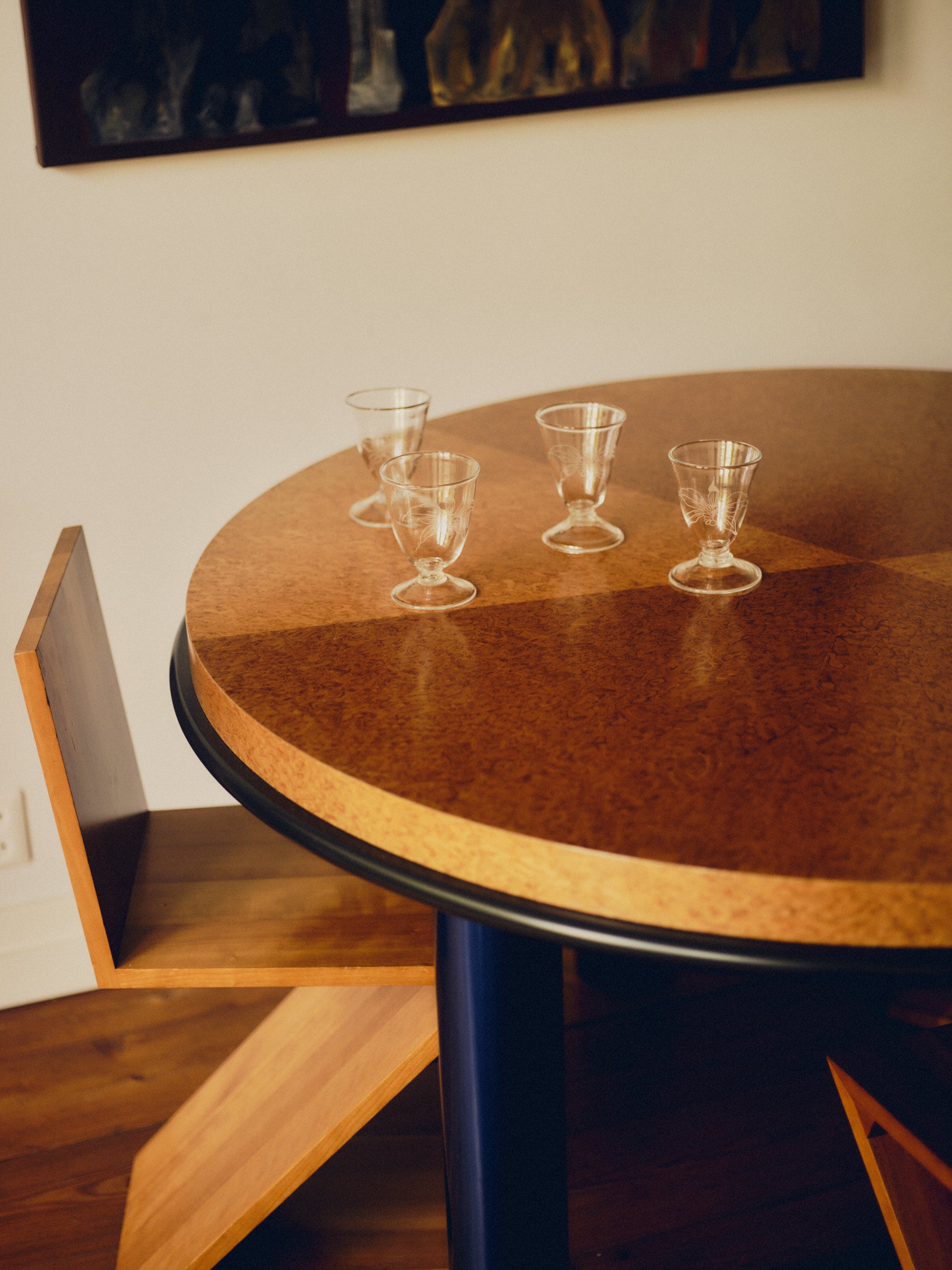 Four wine glasses with engraved flowers by Bastian Bjerge sat on a round, burl wood table with a Zig Zag chair beneath it and a painting hanging above. 