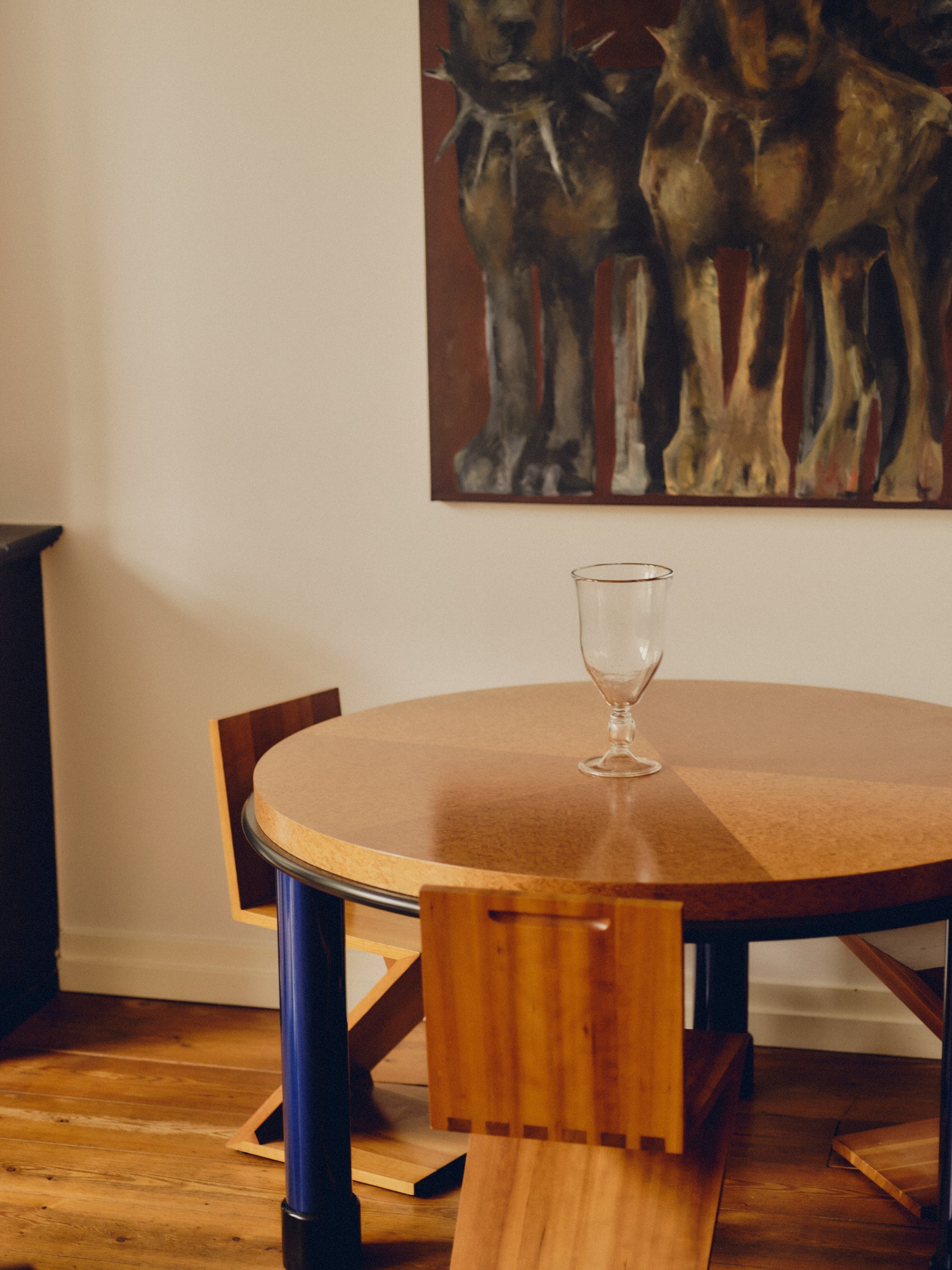 Glass vase with an engraved floral decoration by Bastian Bjerge sat on a burl wood table with three Zig Zag chairs beneath it and a large painting hanging above. 