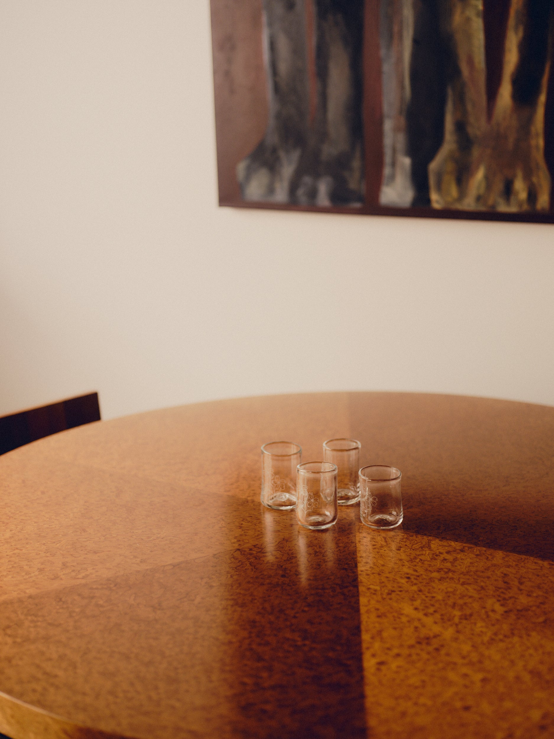 Four drinking glasses with engraved flowers by Bastian Bjerge sat on a round, burl wood table with a painting hang above.