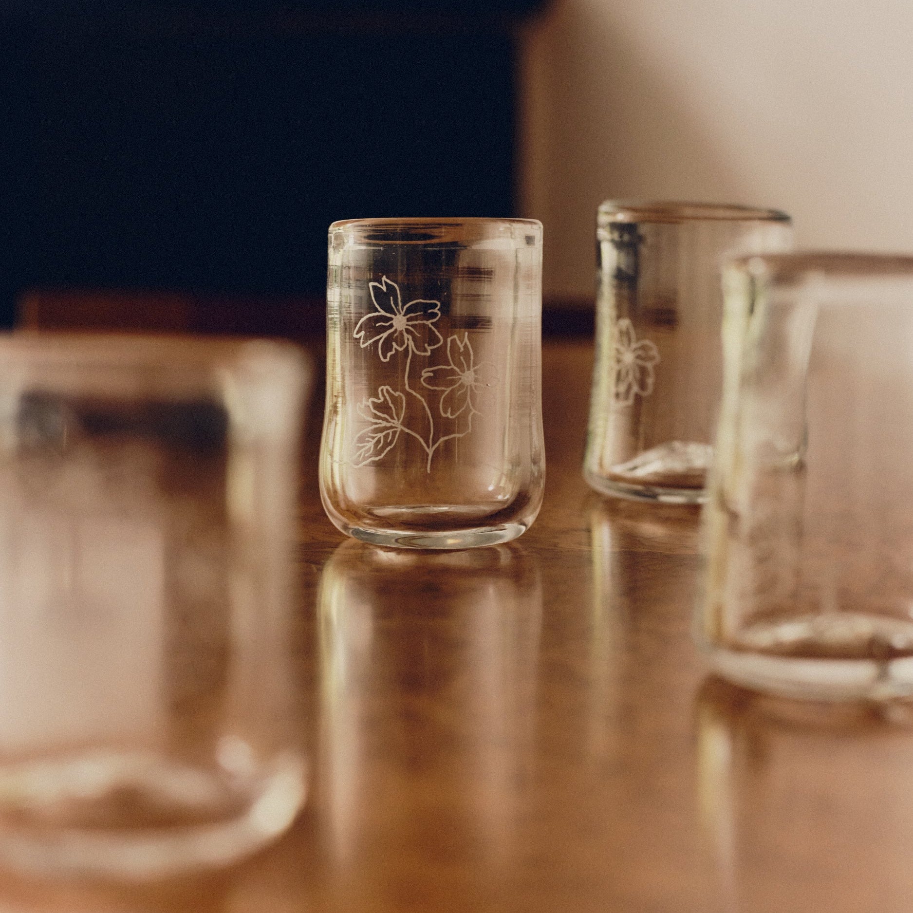 Four drinking glasses with engraved flowers by Bastian Bjerge sat on a burl wood table.