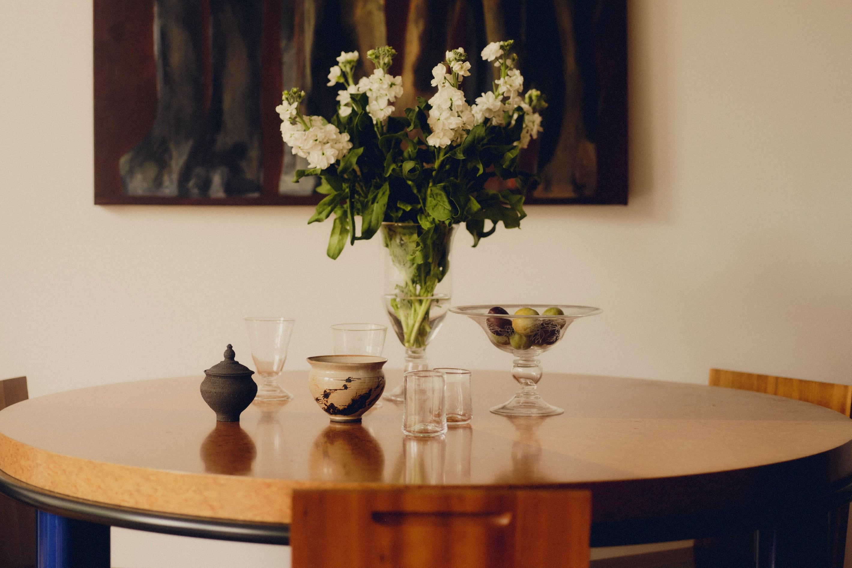 Round wooden table with decorative items including glass cups and a vase of flowers by Bastian Bjerge, with a painting on the wall in the background.