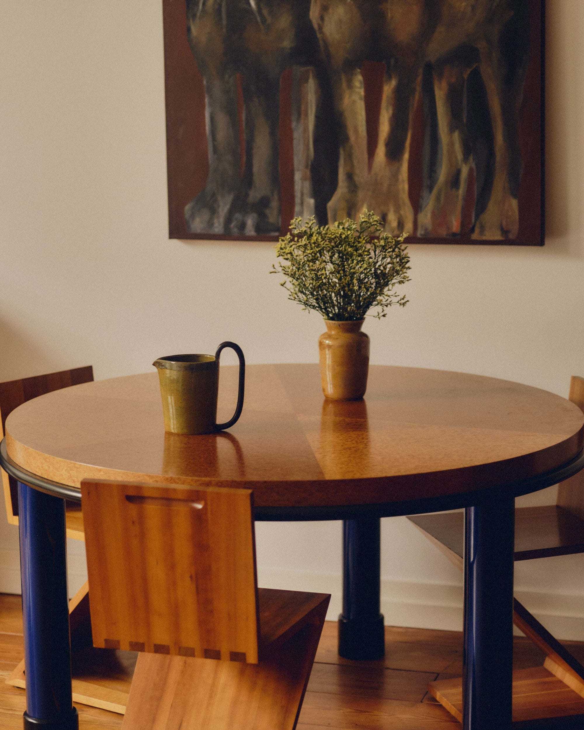 Green ceramic pitcher and ochre glazed ceramic vase holding flowers. Both products by Bira Ceramics are sat on a round burl wood table with blue table legs and Zig Zag chairs beneath it. Wall art hangs above the table.