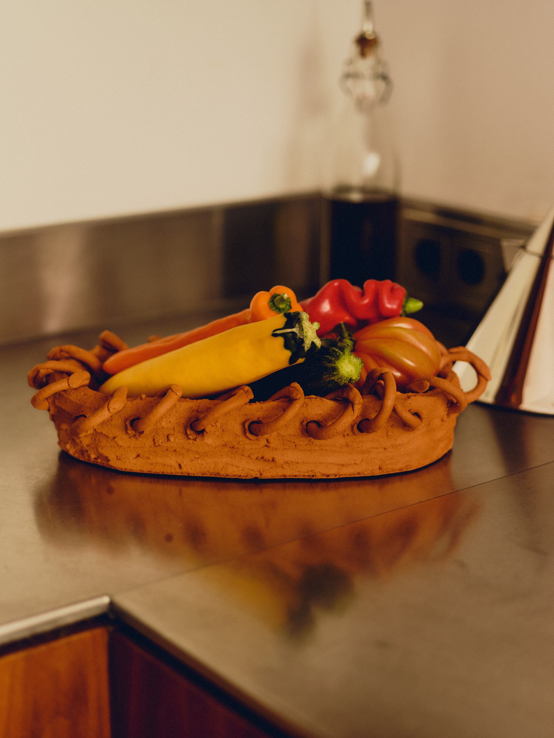 Terra cotta dish with a border of terra cotta rings by May Liok, holding a range of vegetables, sat on a stainless steel kitchen countertop with a blurred glass bottle of oil in the background and the side of a stainless steel kettle.