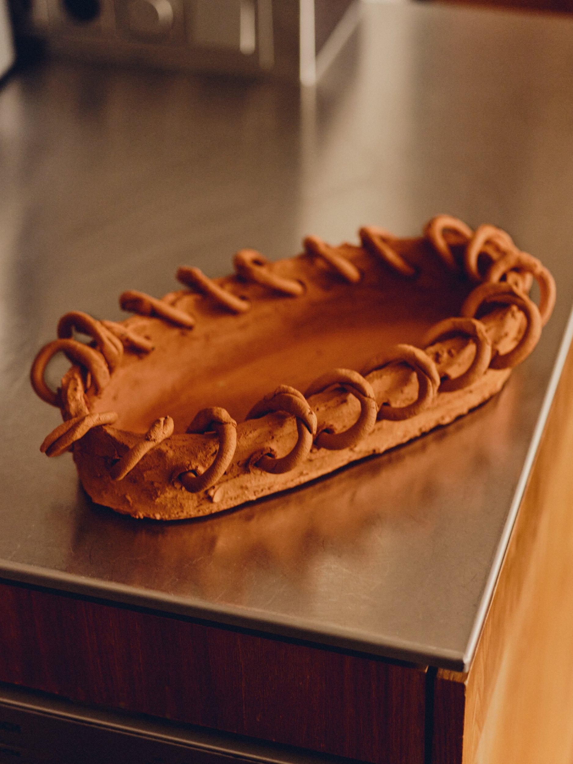 Terra cotta dish with a border of terra cotta rings by May Liok sat on a stainless steel kitchen countertop.