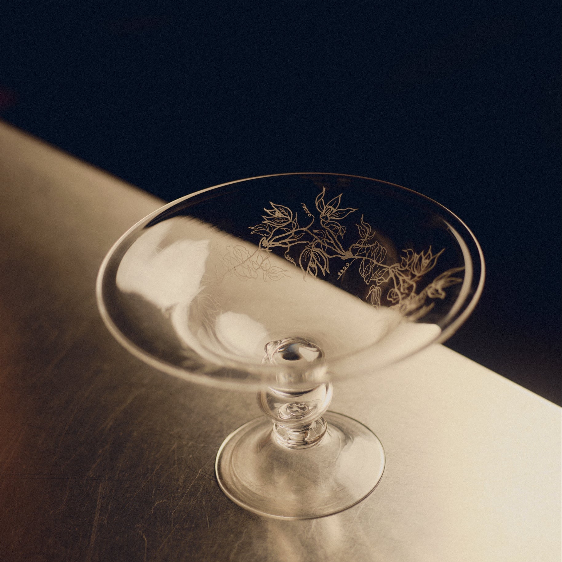 Footed glass bowl with floral engraving by Bastian Bjerge sitting on a stainless steel countertop.