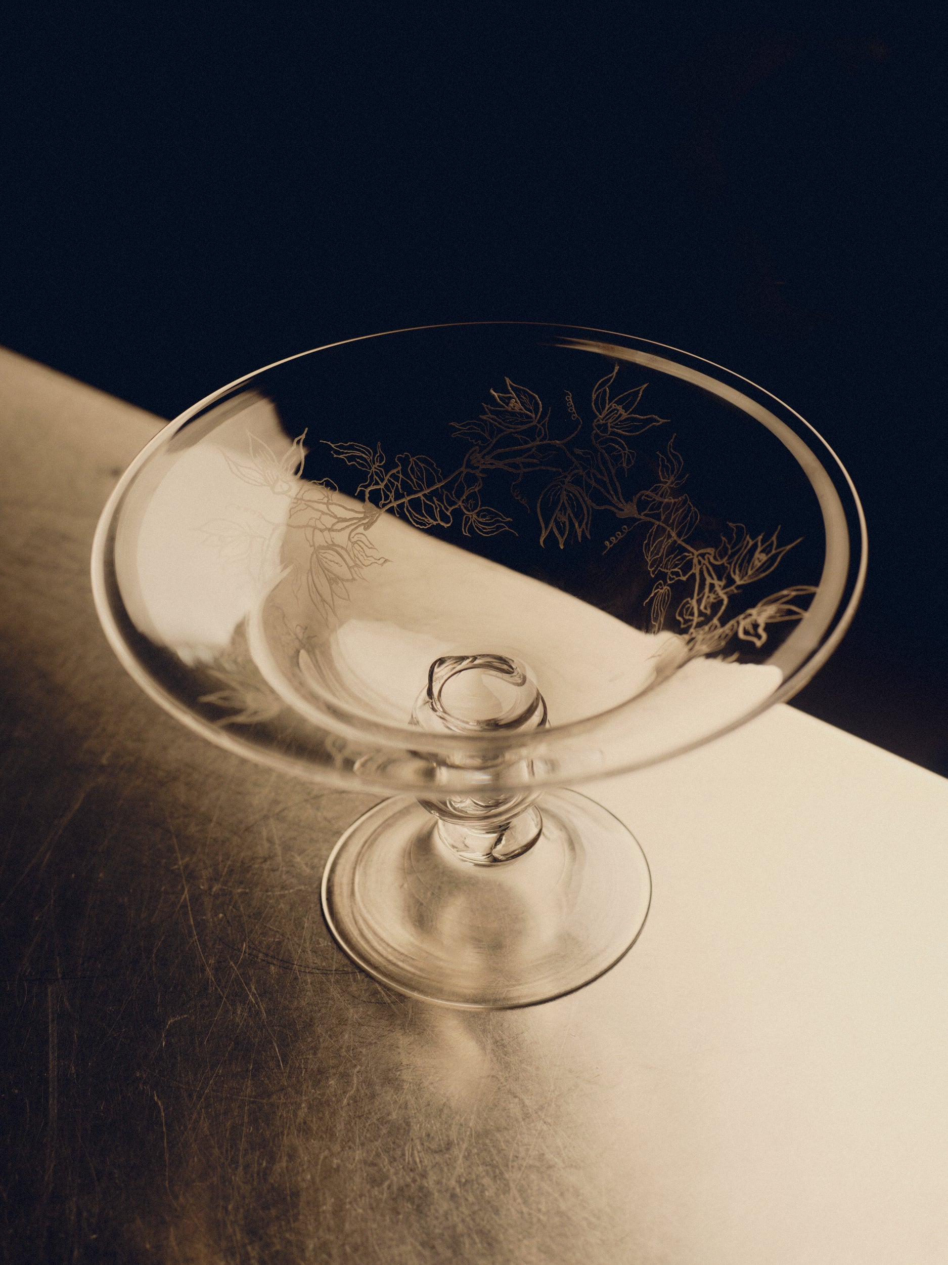 Footed glass bowl with floral engraving by Bastian Bjerge sitting on a stainless steel countertop.