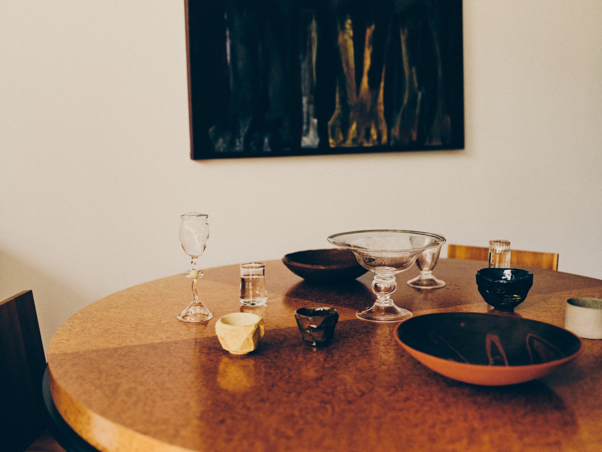 Round wooden table with decorative items including bowls from Klangnapha, Bastian Bjerge and Zeynep Boyan and glasses from Vivian Alarcón and Bastian Bjerge and cups from May Liok, Zeynep Boyan and Klangnapha with a painting on the wall in the background.