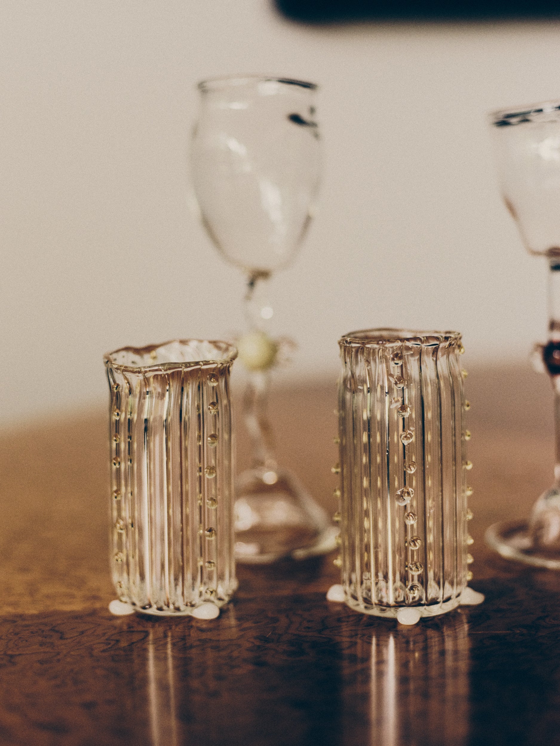 Two mouth-blown, decorative water glasses by Vivian Alarcón on a burl wood table with two wine glasses blurred in the background.