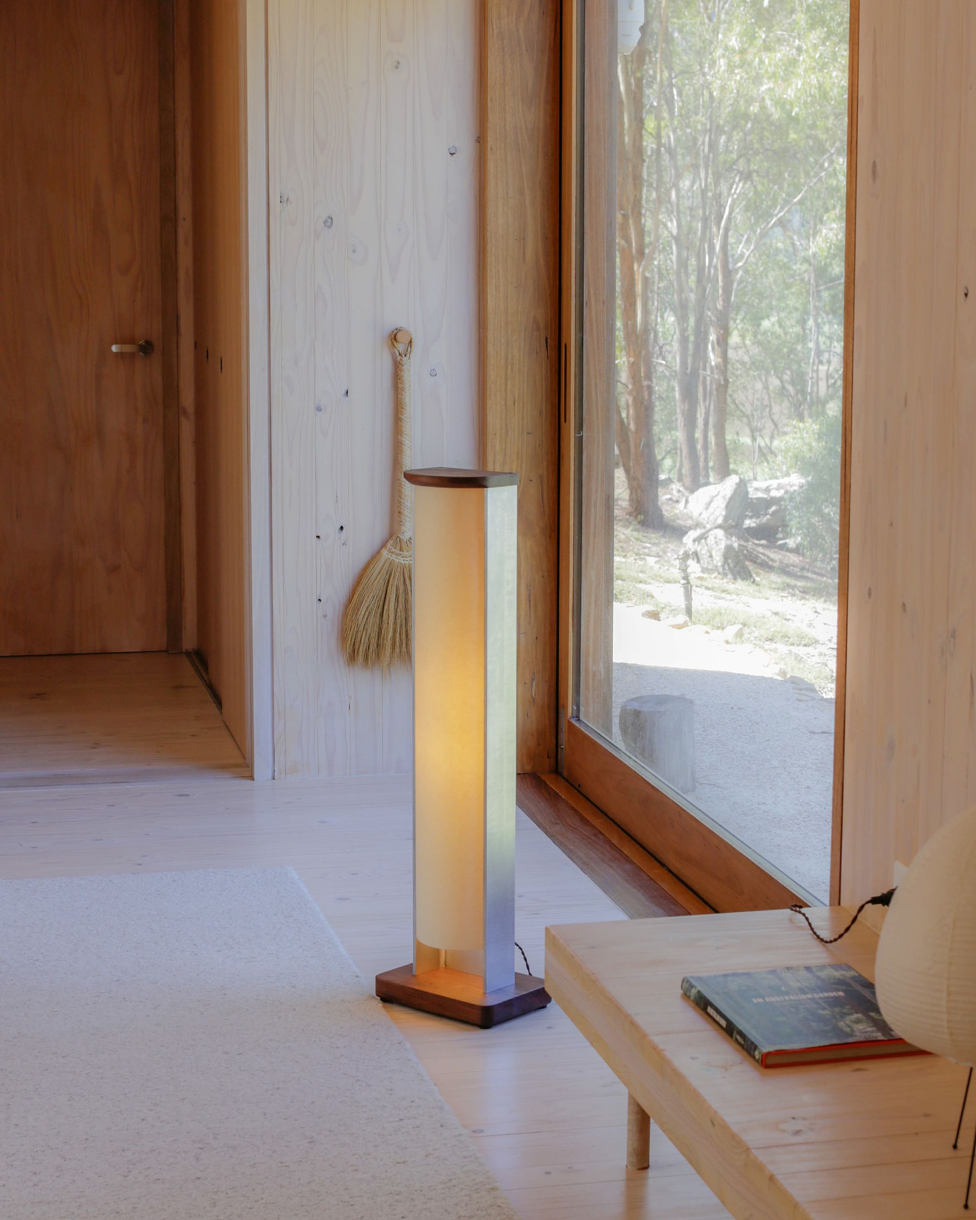 Floor lamp by PARCTA casting a warm glow in a wooden cabin style room. There's woodland through the bright window and a side table beside the lamp.