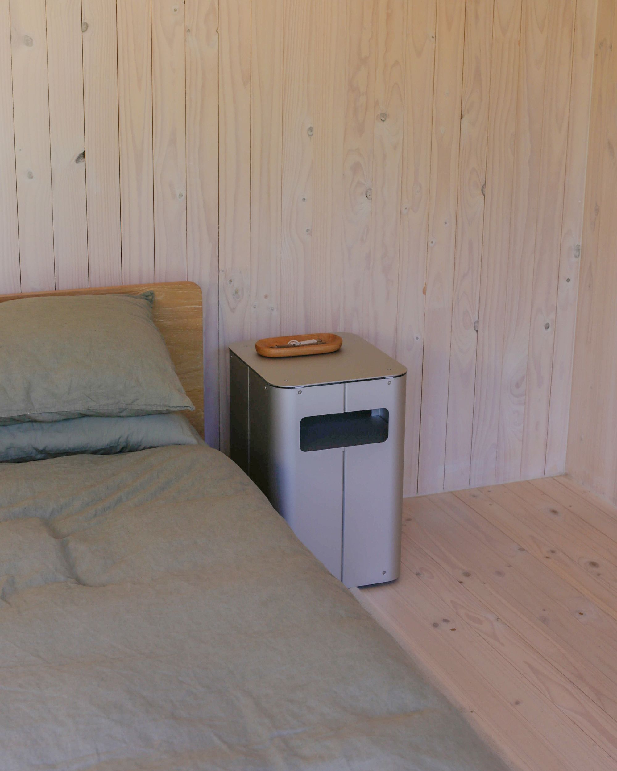 Bedside table by PARCTA placed besides a bed with green linen in a wooden cabin.