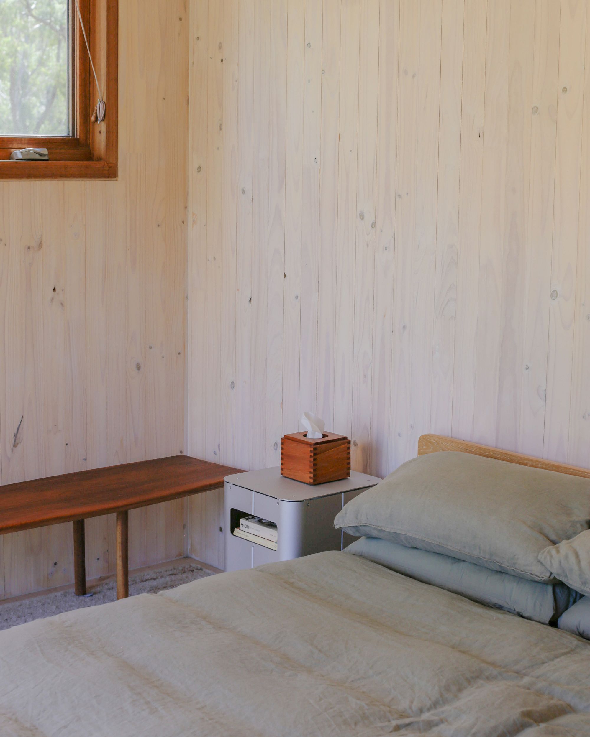Bedside table by PARCTA placed besides a bed with green linen in a wooden cabin. On top of the bedside table is a wooden box of tissues, a bench and a glimpse of a window.