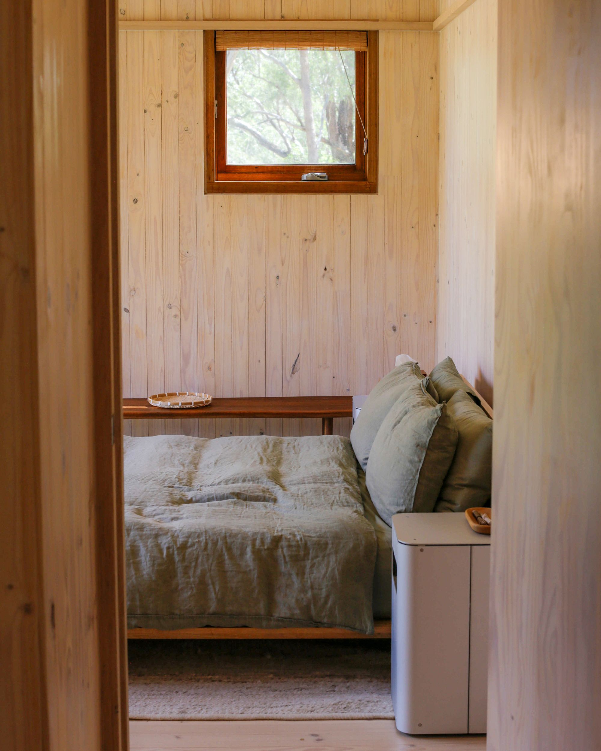 Looking into a wooden cabin bedroom that shows the bedside table by PARCTA. There's a bed with green linen and a small window showing faint woodland outside.