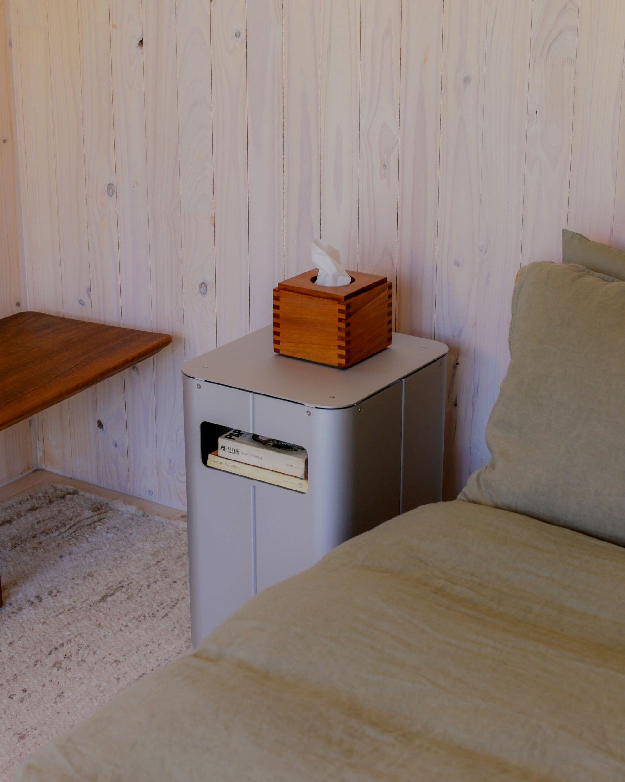 Bedside table by PARCTA placed besides a bed with green linen in a wooden cabin. On top of the bedside table is a wooden box of tissues. 