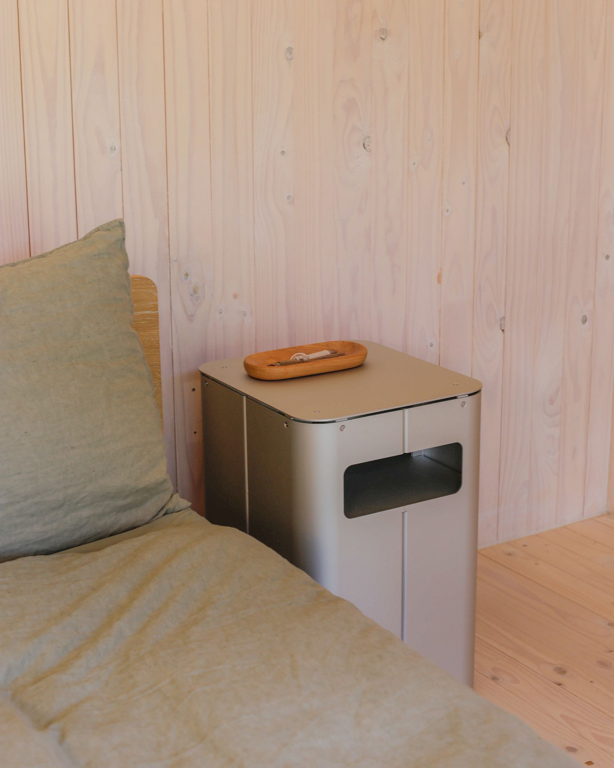Bedside table by PARCTA placed besides a bed with green linen in a wooden cabin.