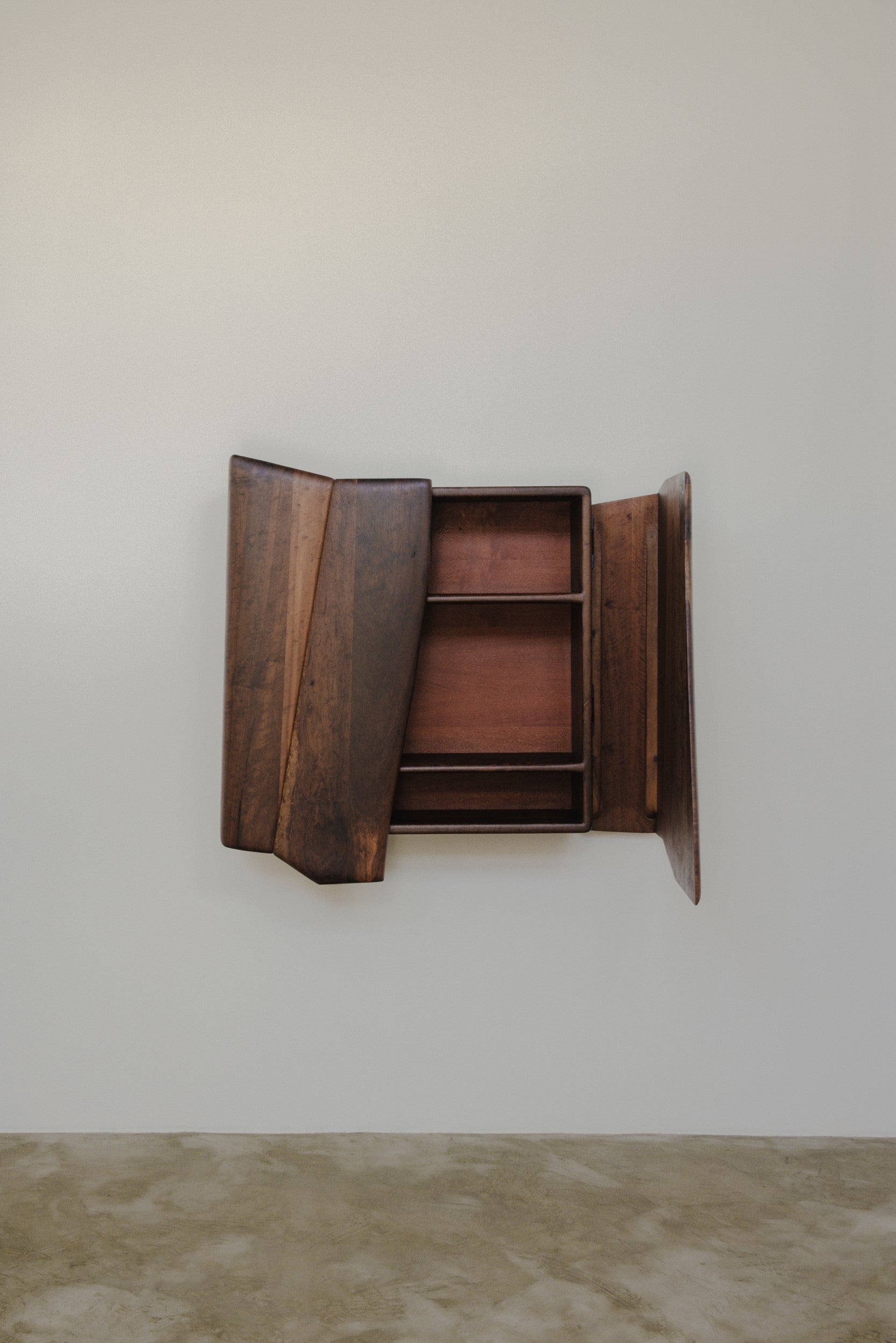Wooden wall mounted cabinet by Estudio Prosa with one door open and one door closed, on a neutral background with concrete floor.