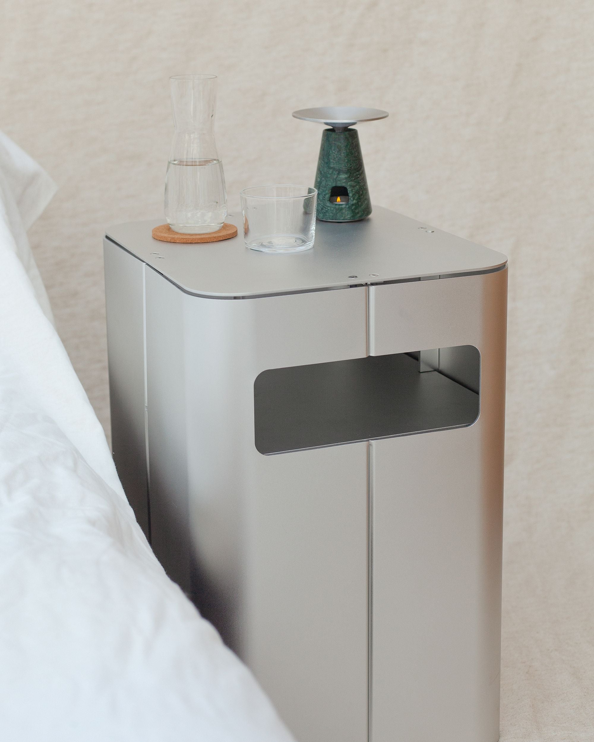 Bedside table by PARCTA placed besides a bed with white linen with a neutral background. There's a small carafe of water with an empty glass and candle.